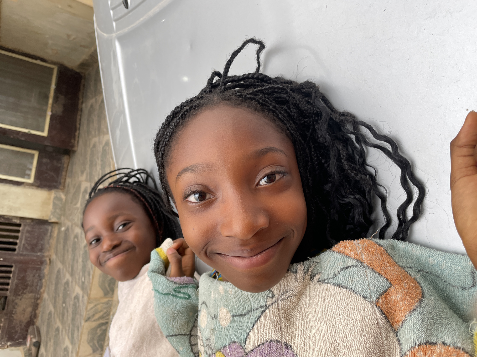 Two young girls are visible in the frame. The primary focus is on the girl closer to the camera, who has her head tilted and is smiling. She has dark braided hair and is wearing a light-colored, patterned sweater. A second girl, also with braided hair, is partially visible to the left. The background includes what appears to be an interior wall and a white or light-colored surface above. The location is indicated as Jos, Nigeria. The exact setting or building is unclear.