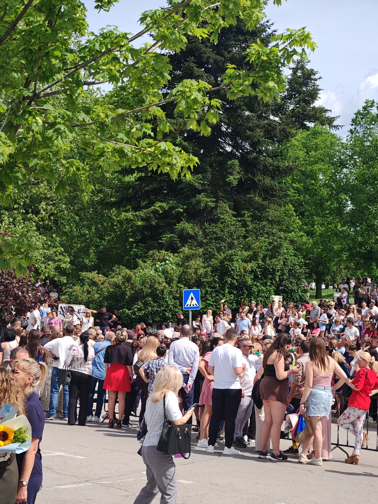The image shows a large crowd of people gathered in what appears to be a public space in the city of Shumen, Bulgaria. The crowd is dense, with people of various ages and ethnicities present. They are standing on an asphalt surface with some visible lane markings. The crowd extends back towards a green area filled with trees and bushes. In the background, behind the crowd, is a blue and white pedestrian crossing sign. The lighting indicates it is daytime, with some areas in shadow due to the trees. The atmosphere seems to be that of an event or gathering of some kind, as opposed to a disorganized mob.