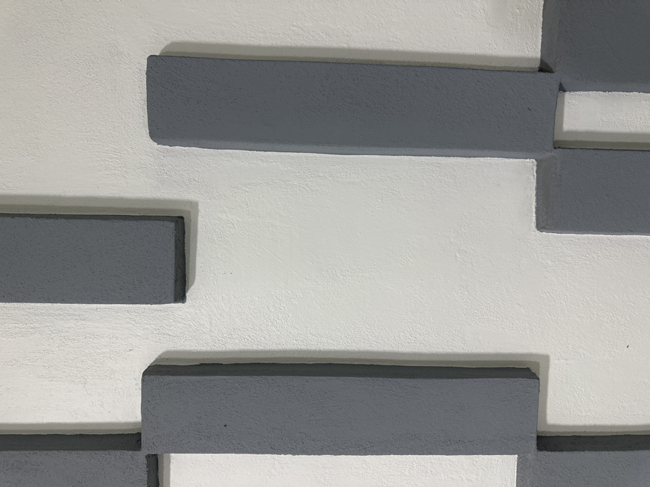 A textured, off-white wall in Mexico City, Mexico, features a geometric arrangement of raised, matte gray rectangular elements. These elements, varying in size and orientation (vertical and horizontal), are affixed to the wall surface, creating a three-dimensional pattern. Shadows are visible along the edges of the raised gray rectangles, indicating their relief against the lighter background. No individuals or discernible objects are present within the frame, and no actions are occurring.