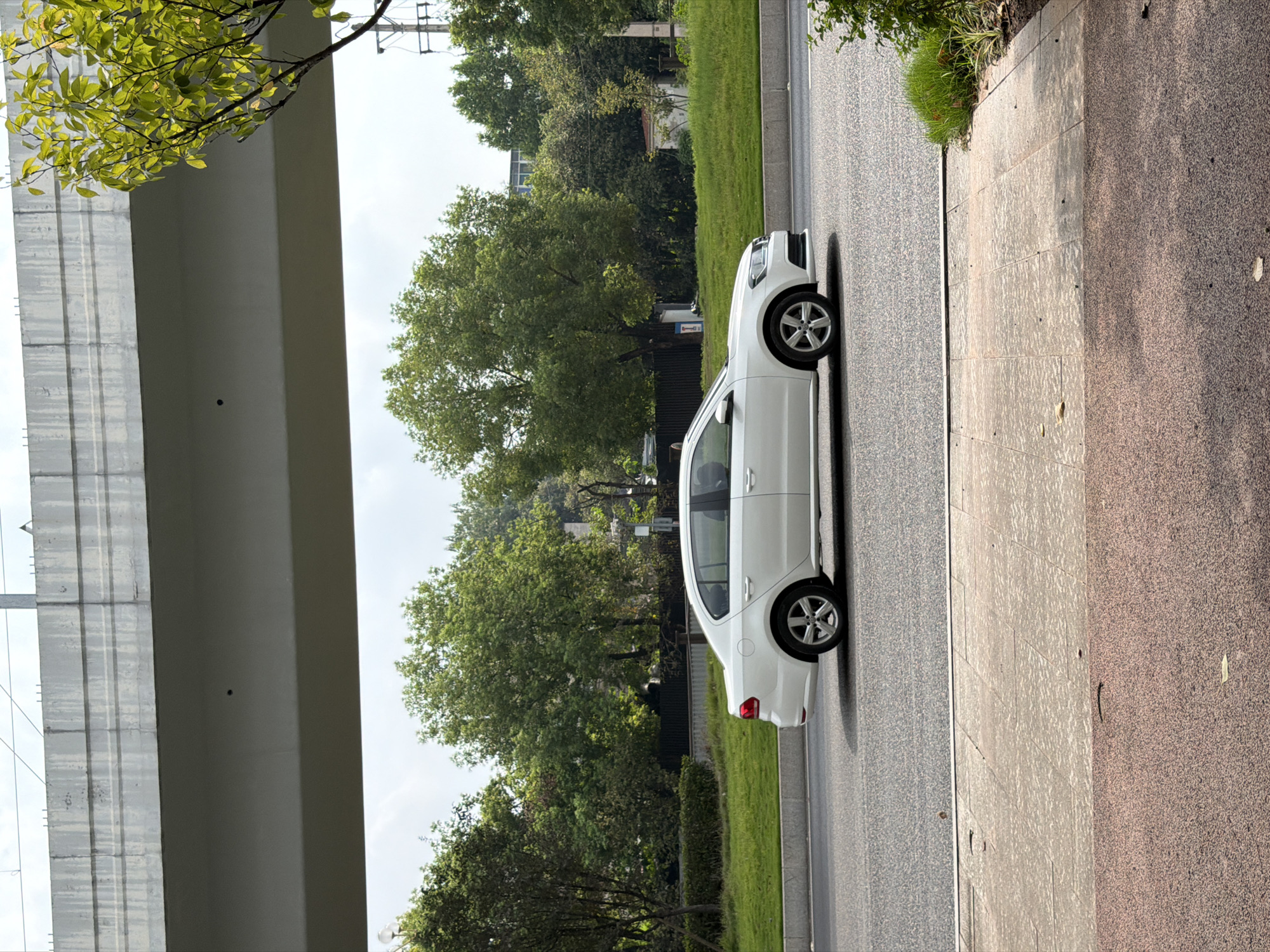 A white sedan is driving on a paved road under an elevated concrete structure. The car is in the right lane, moving from left to right. The elevated structure has parallel lines and supports visible above. Beyond the road, there is a grassy median with trees and some buildings in the background. The sky is overcast. The foreground shows a pedestrian walkway made of stone pavers. The overall scene appears to be in an urban or suburban area of Jiangjin, China.