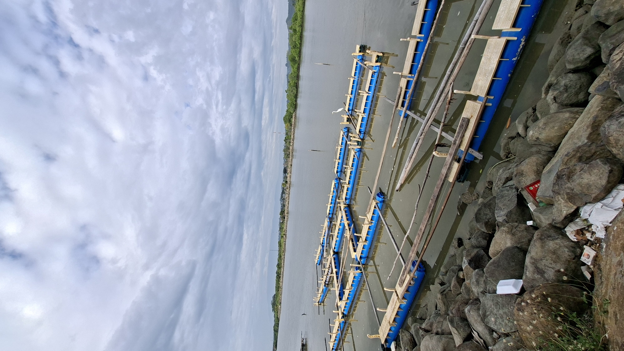 The image depicts a body of water with multiple floating structures, likely fish cages or aquaculture platforms, arranged in rows. These structures are constructed with blue cylindrical pontoons supporting wooden decking. The water is calm, reflecting the overcast sky filled with numerous clouds. The foreground consists of a rocky shoreline with some scattered debris, including plastic waste. In the distance, a green, treed shoreline is visible with some structures and hills beyond. A single white egret stands on one of the floating platforms. This scene is located in Lhokseumawe, Indonesia, a coastal city known for its aquaculture industry.