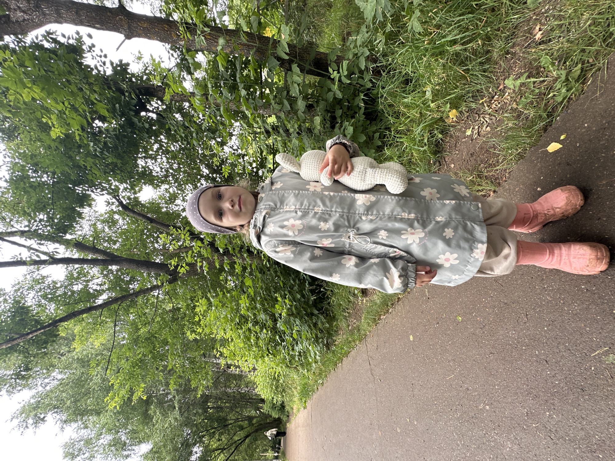 A young child, wearing a grey hat, a light blue floral-patterned jacket, and pink rubber boots, stands on a paved path in an outdoor setting. The child is holding a white knitted stuffed animal. The path is bordered by green grass and trees, with a dense canopy of leaves overhead, suggesting a wooded or park-like environment. The ground appears damp, and there are some fallen leaves on the path, indicating recent rain or damp weather. The overall lighting suggests it could be daytime, possibly overcast given the diffused light. In the background, further down the path, another person is visible in the distance. The image has a casual, candid feel, capturing a moment of a child outdoors, likely on a walk. The location is identified as Chaikovsky, Russia. There is no visible text in the image.