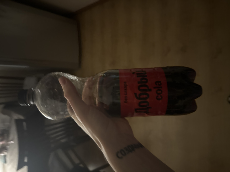A person holds a plastic bottle of Добрый Cola. The bottle is mostly empty, clear plastic near the top, with dark liquid towards the bottom. A red label with white text in Cyrillic is visible. The person's arm, featuring a small tattoo, is visible. The background appears to be a dark room with various objects and furniture blurred.