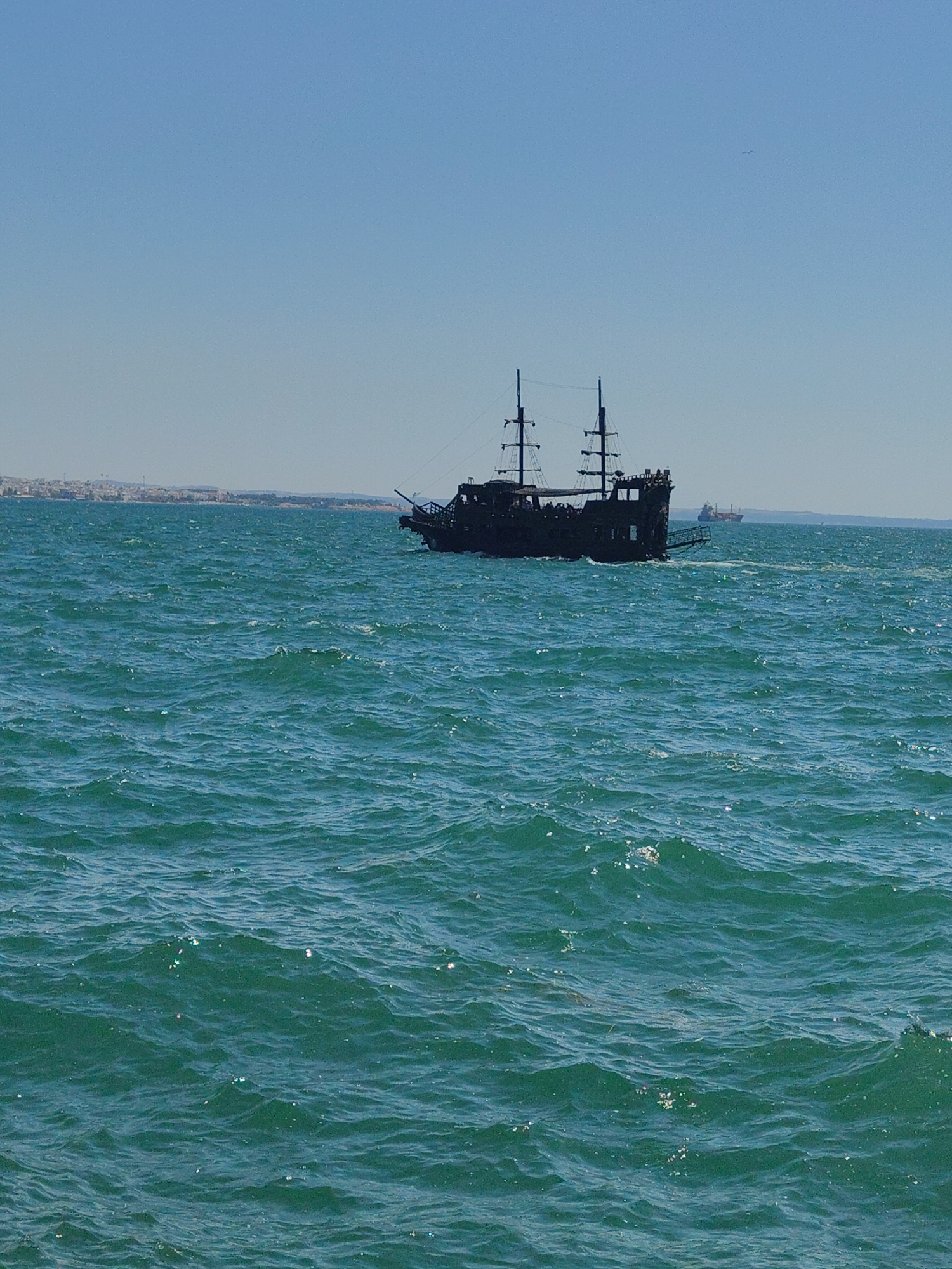A black, three-masted pirate ship sails on a choppy, turquoise sea under a clear blue sky. The ship is silhouetted against the horizon. In the distance, a coastline with buildings is visible. Two other ships, one larger and one smaller, are also present on the horizon. The overall impression is a bright, sunny day at sea.