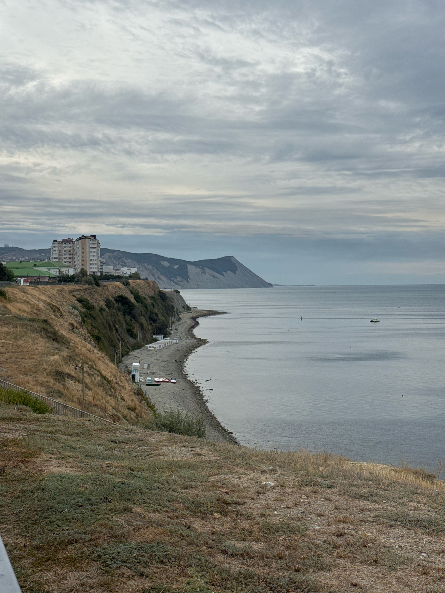 The image captures a panoramic view of a coastal area, likely in Anapa, Russia. The scene is dominated by a body of water, possibly the Black Sea, under a cloudy sky. In the foreground, a grassy embankment slopes down towards a pebble beach. Along the beach, there are several white beach umbrellas and two small boats, one red and one white. Further inland, a steep, dry, grassy cliff rises, topped by several multi-story buildings. The buildings appear to be residential or hotel complexes. In the distance, mountains form a backdrop against the horizon, partially obscured by the overcast sky. A small boat can be seen in the water. The overall impression is one of a somewhat overcast day at a seaside resort.