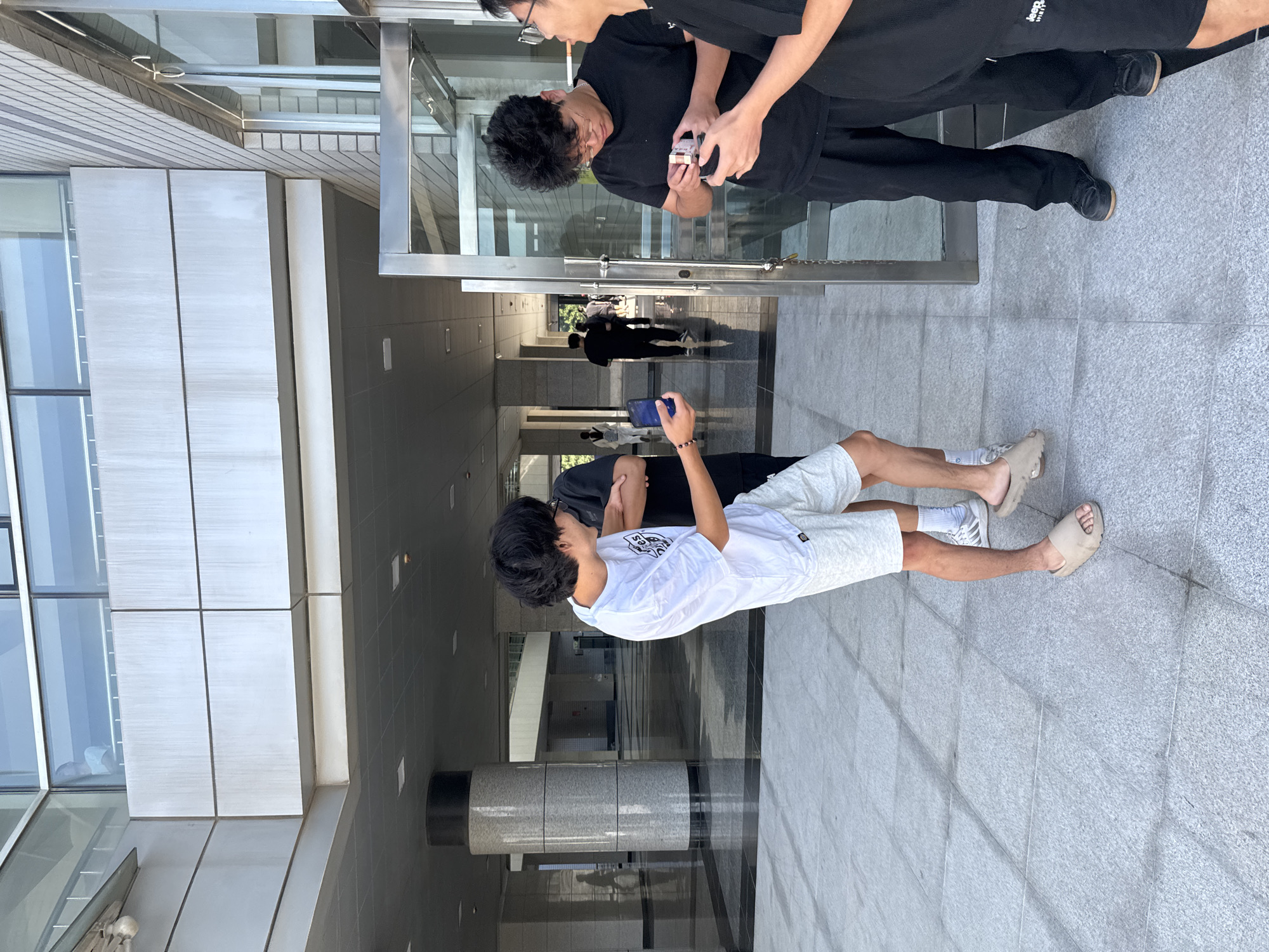 This image depicts a group of young men in an outdoor, urban environment, likely on the grounds of a modern institutional building. The architecture features large columns, sleek metal finishes, and extensive use of glass, suggesting a contemporary design. The flooring is paved with large, light-colored tiles, with darker patterned tiles forming a pathway. Sunlight illuminates the scene, casting shadows and highlighting the textures of the materials.
In the foreground, a young man wearing a white t-shirt and light grey shorts is holding a smartphone and looking towards another individual. He is barefoot and appears to be mid-conversation or engagement. To his right, two other young men are interacting. One, wearing a black t-shirt and black pants, is holding a small object, possibly a pack of cigarettes or a similar item, and examining it closely. The other individual, partially visible on the far right, is wearing black shorts and also appears to be looking at the object.
In the background, other individuals are walking through the covered walkway of the building, suggesting a public or semi-public space. The overall impression is one of casual interaction among friends in a well-maintained, modern urban setting. The city of Guta, China, is a location where such modern architectural styles and public spaces are common, especially in educational or corporate environments.