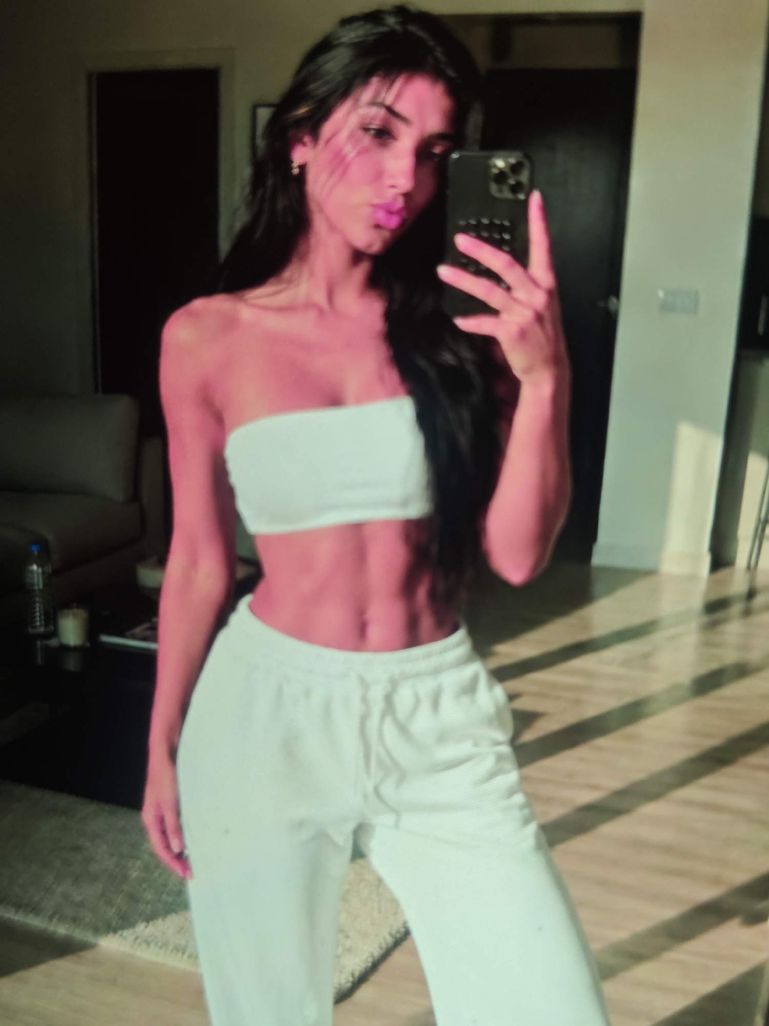 A woman is taking a selfie in a room with sunlight streaming in through the blinds. She has long, dark hair and is wearing a white bandeau top and white sweatpants. She is holding a smartphone in her right hand and making a pouty face. In the background, there is a couch, a table with a bottle and a candle, a doorway, and a wall with a light switch. The floor is made of wood and there is a rug.
