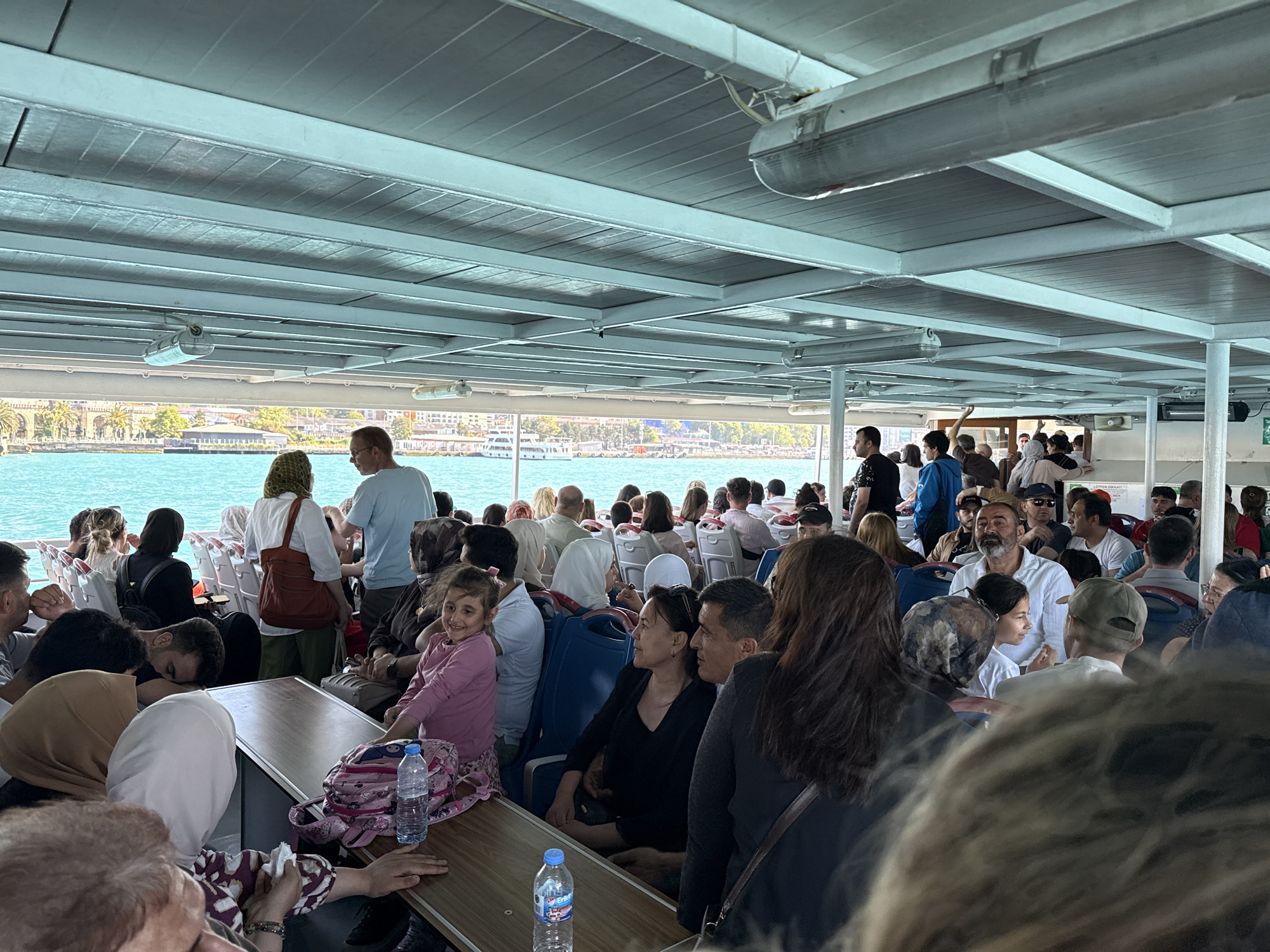 The image shows a crowded ferry in Beyoğlu, Turkey. Passengers of various ages are seated and standing, filling the interior space. A few are looking out the windows at the water and distant coastline. Bottles of water and bags are on the tables. The ceiling of the ferry is visible, with overhead lighting. Outside the ferry, the water is turquoise, and land with buildings and trees is in the background.