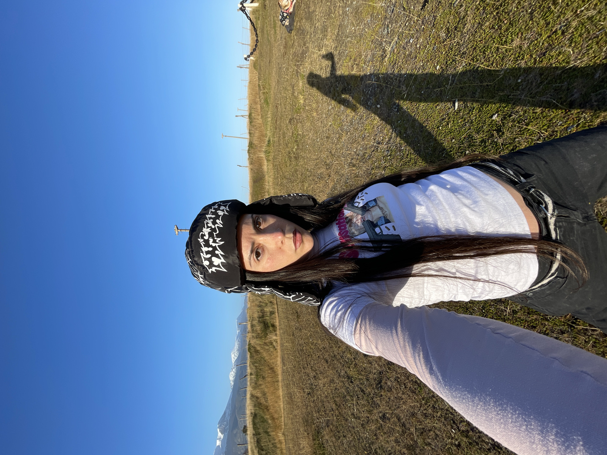 A woman wearing a black and white patterned head covering, a white t-shirt, and dark jeans is lying on a grassy field. The field contains dry grass and green patches of vegetation. A shadow of a person is cast on the grass in the background. Power lines are visible in the distance. In the far distance, snow-capped mountains are visible against a clear blue sky. The image was taken in Ushuaia, Argentina.