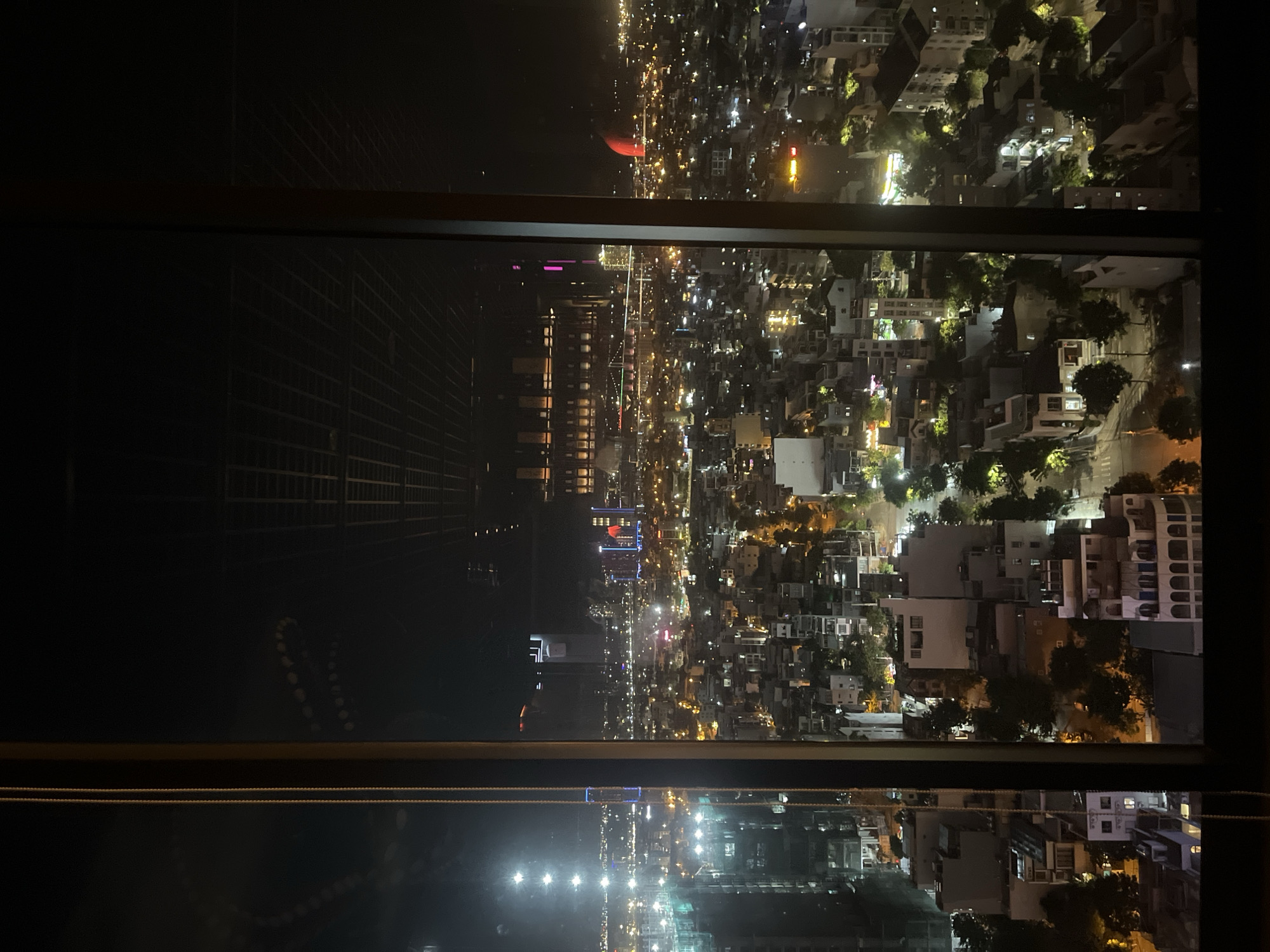 This image captures a nighttime aerial view of a cityscape, likely Đà Nẵng, Vietnam, seen through a window. The scene is divided horizontally by a frame, with the top portion displaying the city lights and the bottom portion showing a reflection of the scene below.
In the upper part of the image, the city is illuminated by countless lights from buildings and streets, creating a vibrant, sprawling urban landscape. The density of buildings suggests a well-developed urban area. Scattered among the structures, the warm glow of streetlights and the lights from within buildings paint a picture of a city alive at night. A notable detail in the distance is a large, bright red object, possibly a hot air balloon or an illuminated structure, standing out against the dark sky. In the lower portion of the image, a detailed reflection of the city and the window frame is visible on a dark, possibly glass or water surface. The reflection mirrors the cityscape above, including the vibrant lights and the structural elements of the buildings, albeit slightly distorted and with a darker tone.
The time of day is clearly night, given the darkness of the sky and the prevalence of artificial lighting in the city. The weather appears clear, as the lights are sharp and there are no visible signs of fog or heavy rain. The setting is an elevated vantage point, such as a high-rise building or a hotel room, offering a panoramic view of the city. The overall mood evoked is one of urban energy and vastness, with the reflections adding a layer of depth and visual interest. There is no visible text in the image.