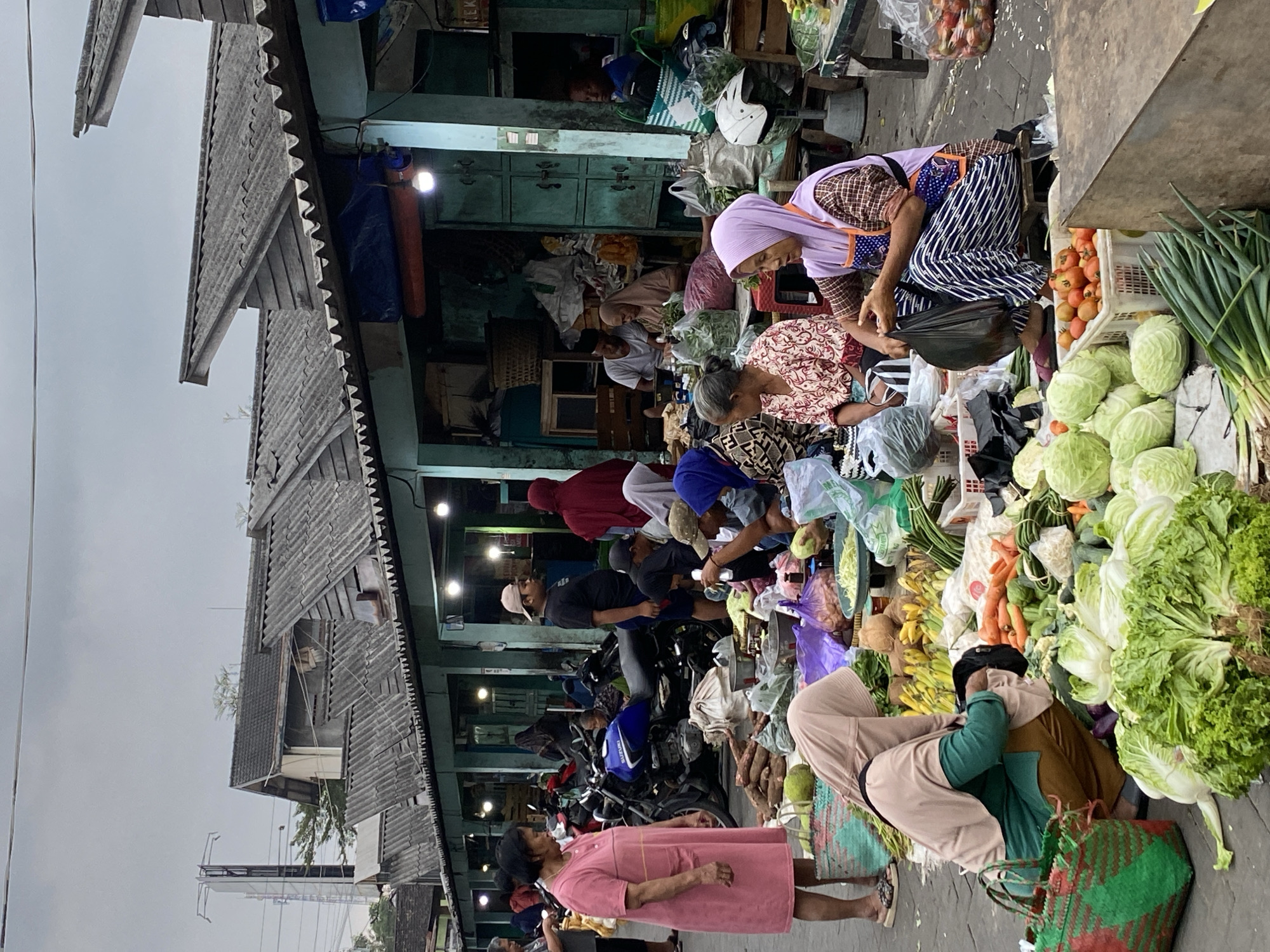 This image depicts an outdoor market scene in Ambarketawang, Indonesia. The market stalls are arranged along a pathway, with covered structures providing shelter overhead. The ground is paved and wet in places, suggesting recent rain or cleaning. A variety of fresh produce, including cabbages, carrots, bananas, and tomatoes, are displayed for sale. Several people, predominantly women, are seen attending to the stalls, interacting with customers, or arranging goods. Some are wearing headscarves and traditional Indonesian clothing. Motorcycles are parked in the background, indicating transportation common in the area. The overall atmosphere suggests a bustling local marketplace.