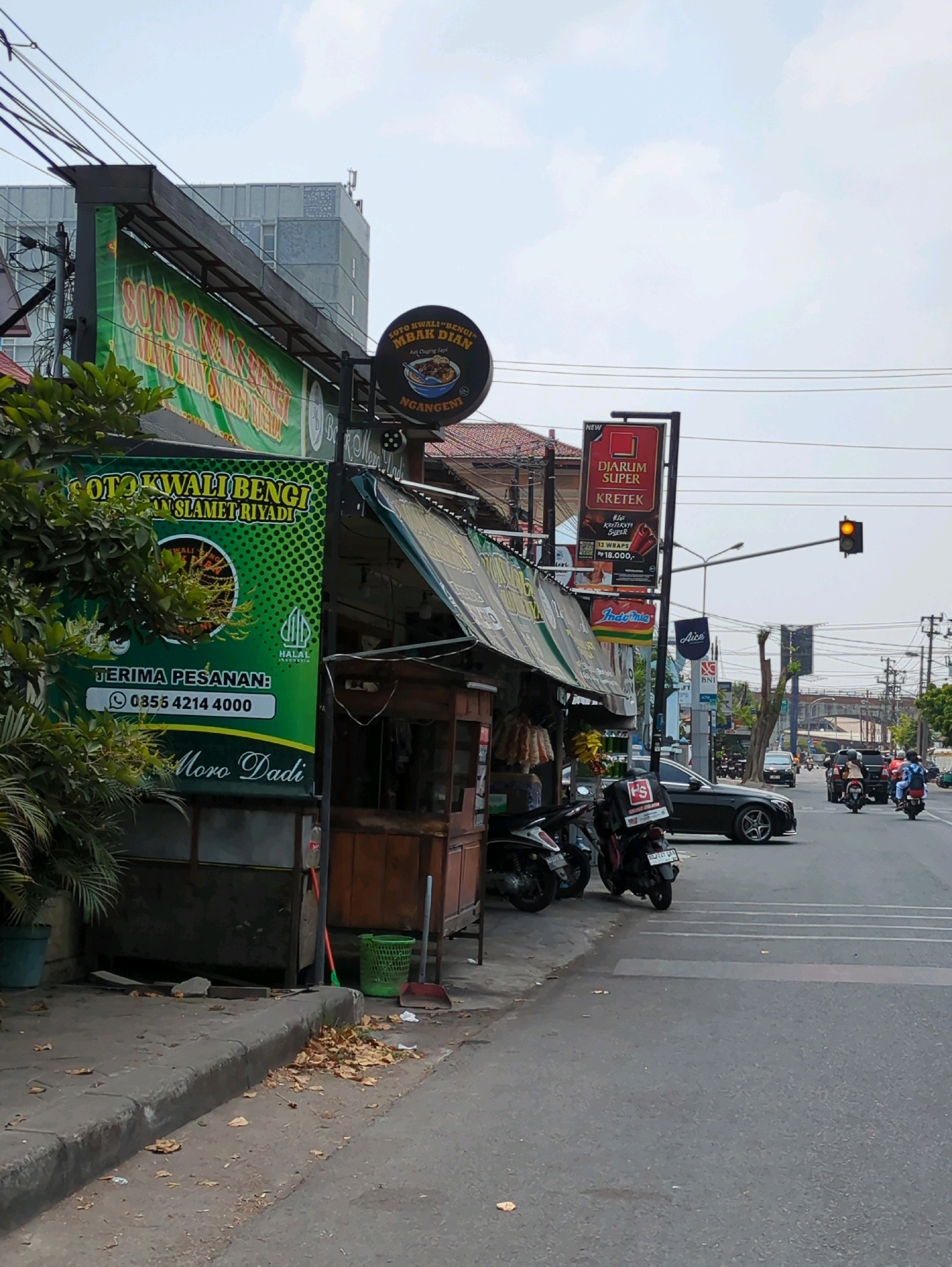 The image shows a street scene in Surakarta, Indonesia, with a focus on a roadside food stall and surrounding environment. On the left, a large green banner advertises "SOTO KWALIBENGIM SLAMET RIYADI" with a phone number for orders, "0855 4214 4000", and the words "Moro Dadi". Above the banner, a smaller black circular sign displays "SOTO KWALIBENGIM MBAK DIAN NGANGENI" along with an illustration of a bowl of food. The food stall itself has a wooden structure with a corrugated metal roof and appears to be open for business, with goods hanging from the side.
In front of the stall, several motorcycles are parked. A black Mercedes-Benz sedan is driving on the right side of the road, and other vehicles, including motorcycles and cars, are visible further down the street. Traffic lights are present, with one showing a yellow light. The environment appears to be a typical urban street with overhead electrical wires, streetlights, and buildings in the background. Fallen leaves are scattered on the sidewalk and street. The sky is overcast.