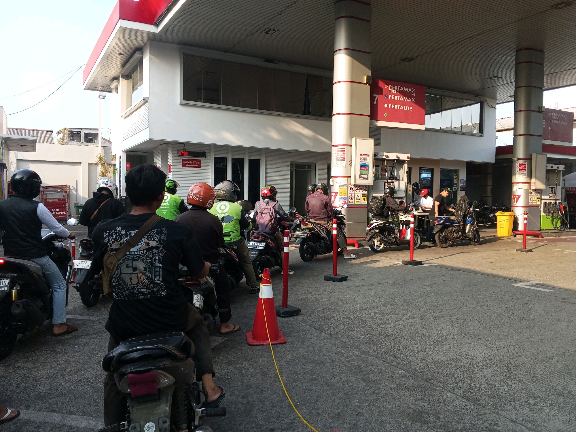 The image depicts a gas station in South Jakarta, Indonesia, on a sunny day. Several people on motorcycles are lined up, seemingly waiting to refuel. The gas station features prominent red and white branding, with signs indicating different fuel types like "PERTAMAX TURBO" and "PERTAMAX". A digital clock displays "07:09". People are dressed casually, some wearing helmets. The environment appears to be a typical urban setting with a covered forecourt and paved ground. Various objects such as traffic cones, fuel pumps, and a yellow trash bin are visible.