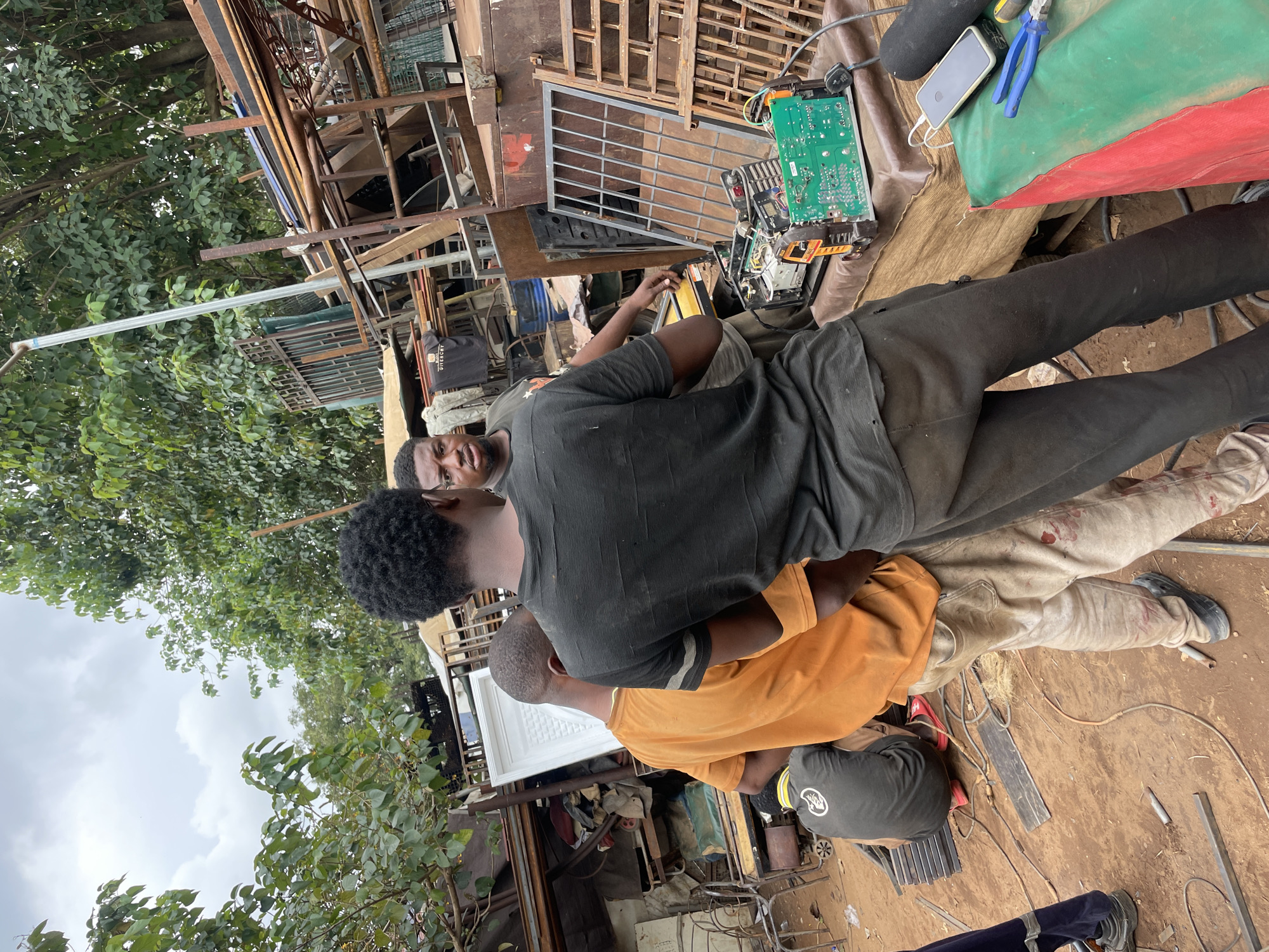 The image depicts a group of three men in what appears to be an outdoor workshop or junkyard setting, likely in Abuja, Nigeria. The scene is cluttered with various metal scraps, construction materials, and what looks like electronic components.
In the foreground, two men are partially visible. One man, wearing a black t-shirt and dark pants, is looking directly at the camera. Another man, wearing an orange shirt, is partially obscured behind him, facing away. A third man, wearing a dark grey t-shirt with a white logo, is lying down or sitting in the background, facing a piece of electronic equipment. This equipment, with a green circuit board, is laid out on the ground, along with a smartphone and a pair of blue pliers.
The overall atmosphere suggests a working environment, possibly involved in electronics repair or salvage. The presence of tools and materials points towards some form of manual labor or technical work. The lighting suggests it could be daytime, with overcast or cloudy skies. The background reveals more scrap metal and foliage, reinforcing the impression of an outdoor, industrial or semi-industrial location. There is no discernible text in the image.