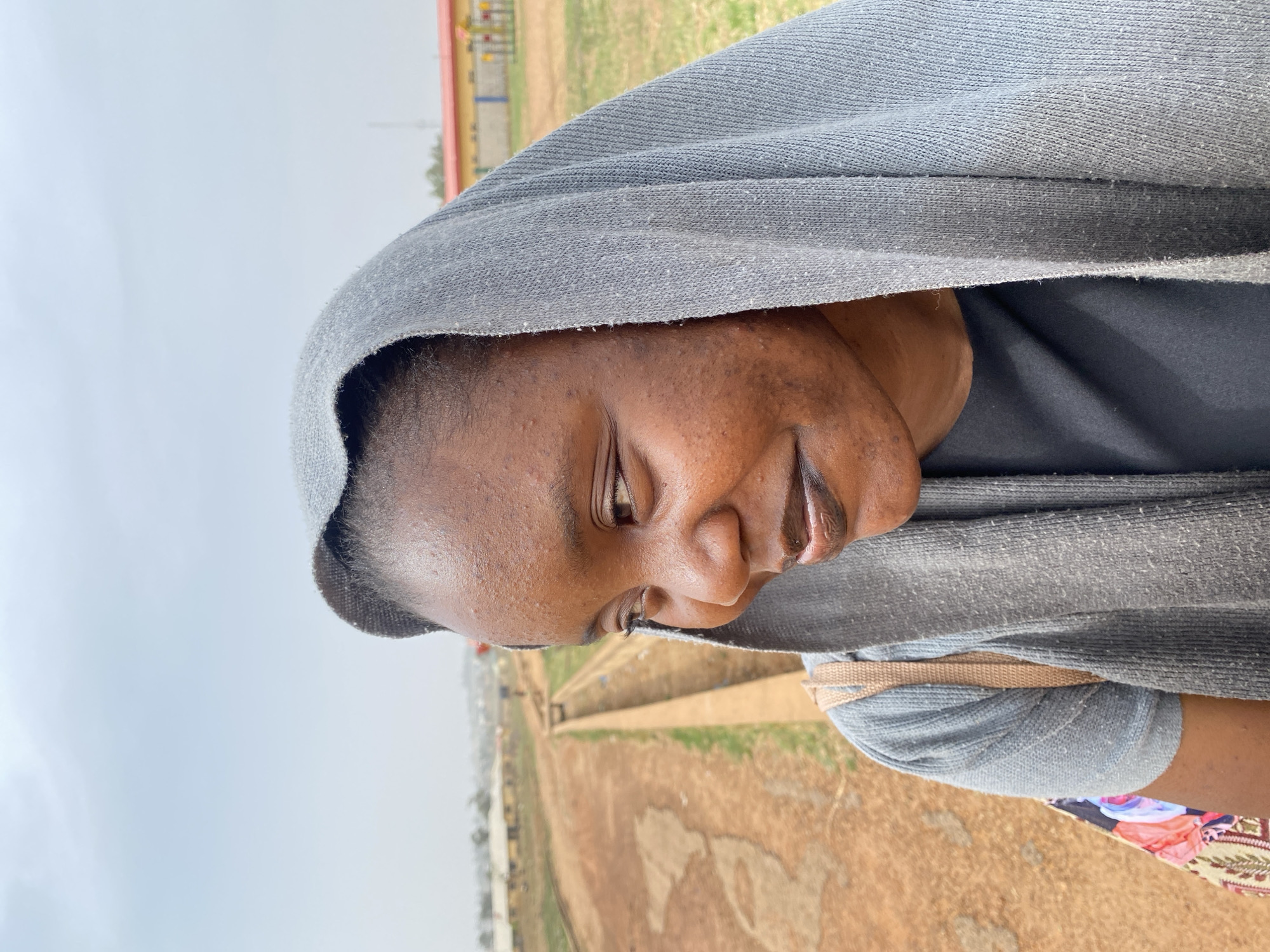 The image, taken in Vom, Nigeria, features a person, likely a woman, captured from the chest up, positioned towards the right side of the frame. She has dark skin and is wearing a light grey head covering or shawl that drapes over her head and shoulders. Underneath, she wears a dark-colored top. Her gaze is directed downwards and slightly to her right, and she has a gentle, subtle smile on her lips. A beige strap, possibly from a bag, is visible over her right shoulder, and a small portion of a colorful, patterned fabric, possibly a skirt or another garment, can be seen near her right arm.
The background depicts an outdoor setting under a bright, overcast, or hazy sky. The landscape is characterized by reddish-brown earth and sparse patches of green grass. A distinct path or channel runs through the middle ground. In the distance, indistinct structures with red and yellow components, possibly playground equipment or buildings, are visible among sparse vegetation. The overall atmosphere suggests a calm, daytime moment in a dry, open environment. No text is visible in the image.