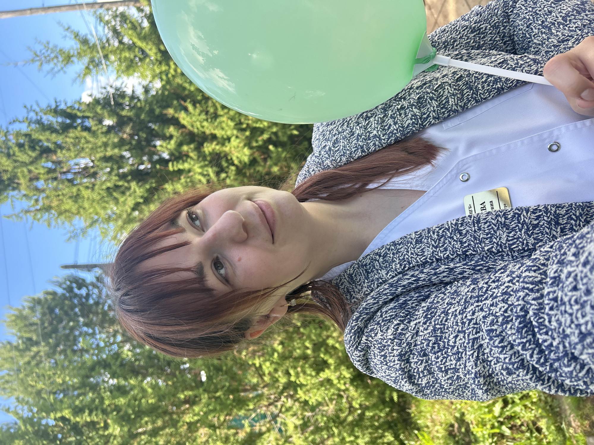 A young woman with auburn hair and bangs, wearing a white shirt and a navy blue knit sweater, holds a pale green balloon. She wears a name tag. The background shows green trees and a blue sky. The photograph may have been taken outside in Фоки, Russia, though this cannot be confirmed.