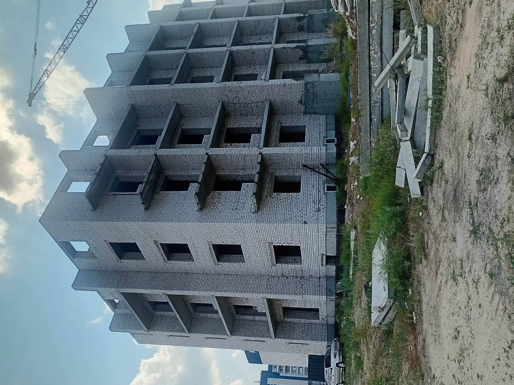 An unfinished multi-story building constructed of white bricks is visible, with concrete balconies and open window frames. A construction crane stands in the background, suggesting ongoing work. In the foreground, stacks of concrete slabs and debris are scattered on the ground. A silver car is parked to the left of the building, and a tall blue and white building is partially visible behind it. The scene is set against a cloudy sky. This image was taken in the city of Semey, Kazakhstan.