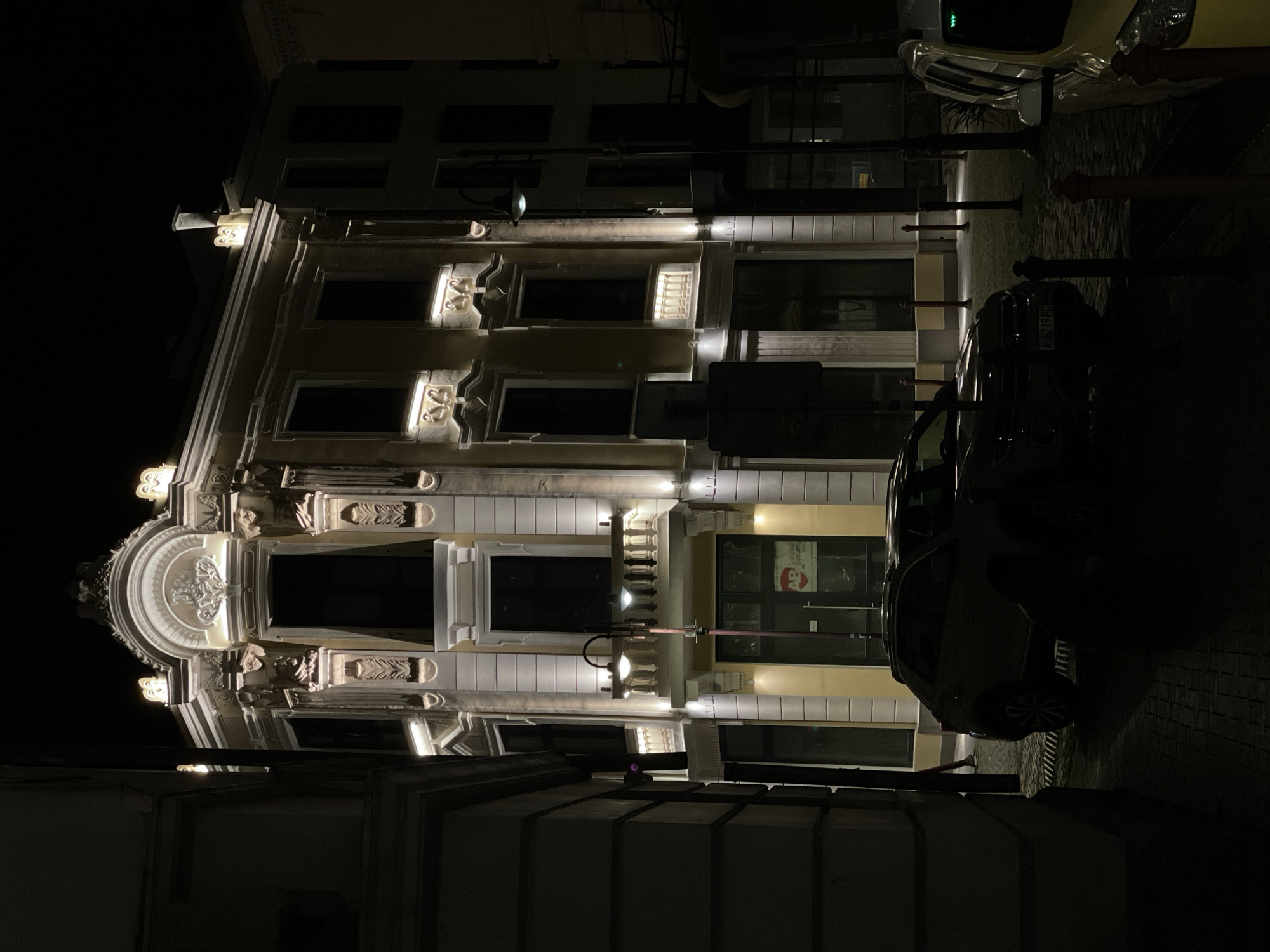 The image shows a building at night in Burgas, Bulgaria. The building has an ornate facade with detailed carvings and is illuminated by spotlights. A car is parked in front of the building. There are street lights and bollards lining the sidewalk. The buildings across the street are also visible in the darkness.