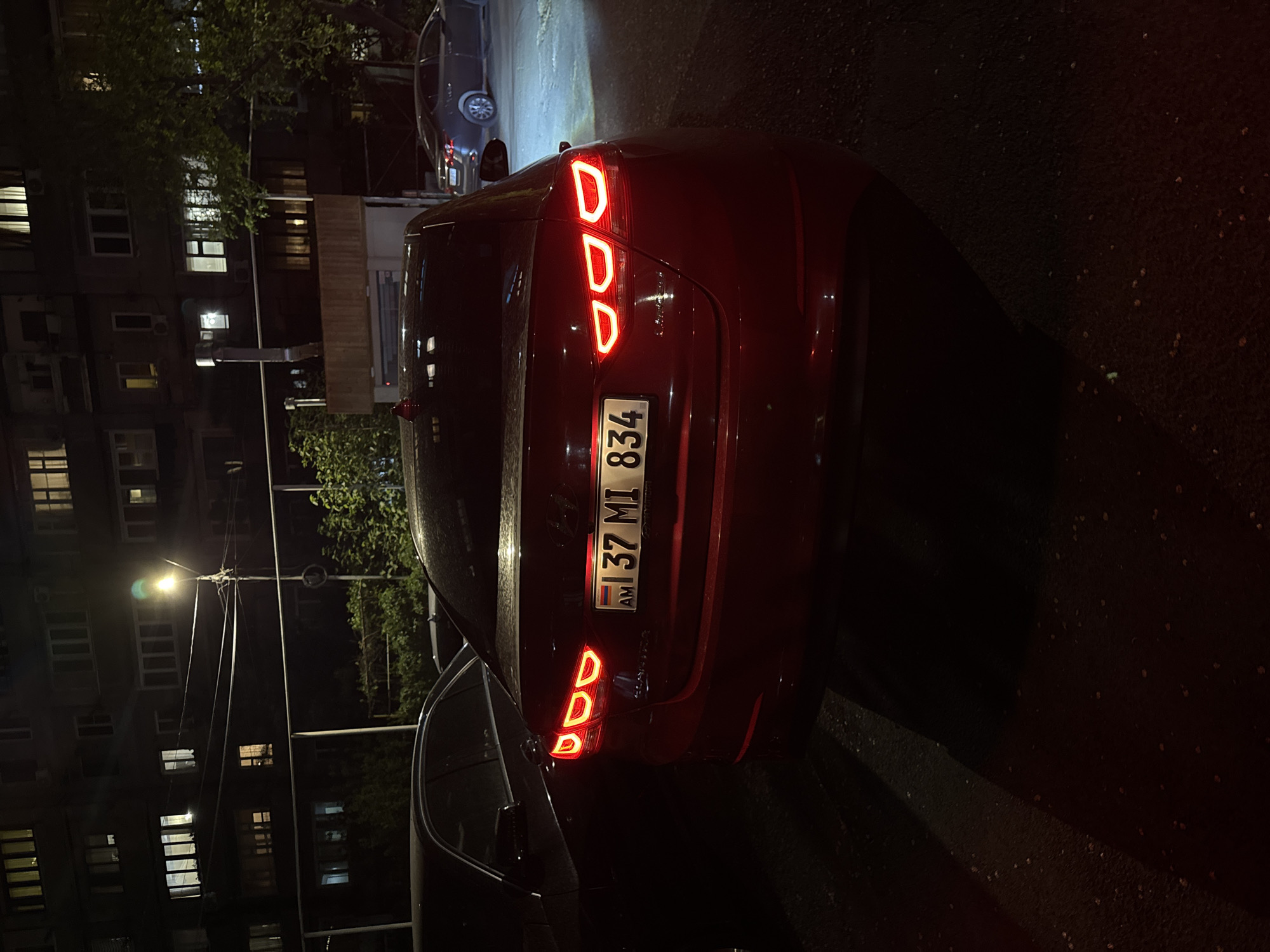 The image shows the rear of a maroon car parked on a street in Yerevan, Armenia. The license plate reads "AM 37 MI 834". The car's taillights are illuminated. The street appears to be in a residential area with apartment buildings visible in the background, illuminated by streetlights. Other vehicles are parked nearby. The scene is captured at night.