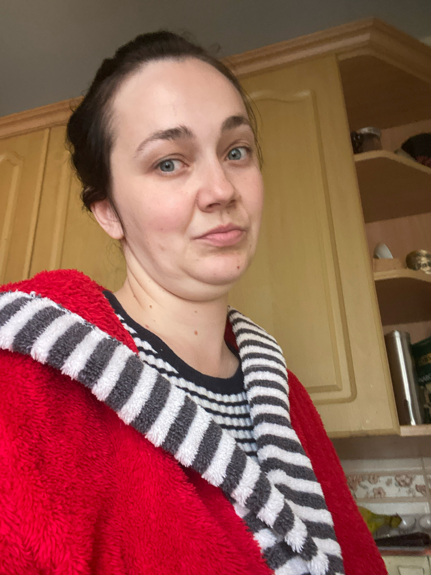 A woman with dark hair pulled back is looking at the camera with a skeptical expression. She is wearing a red robe with a striped collar and a striped shirt underneath. Her chin is slightly tucked, creating a double chin. Behind her, a light wood kitchen cabinet is visible, with shelves containing various items. The background suggests an indoor setting, likely a kitchen.