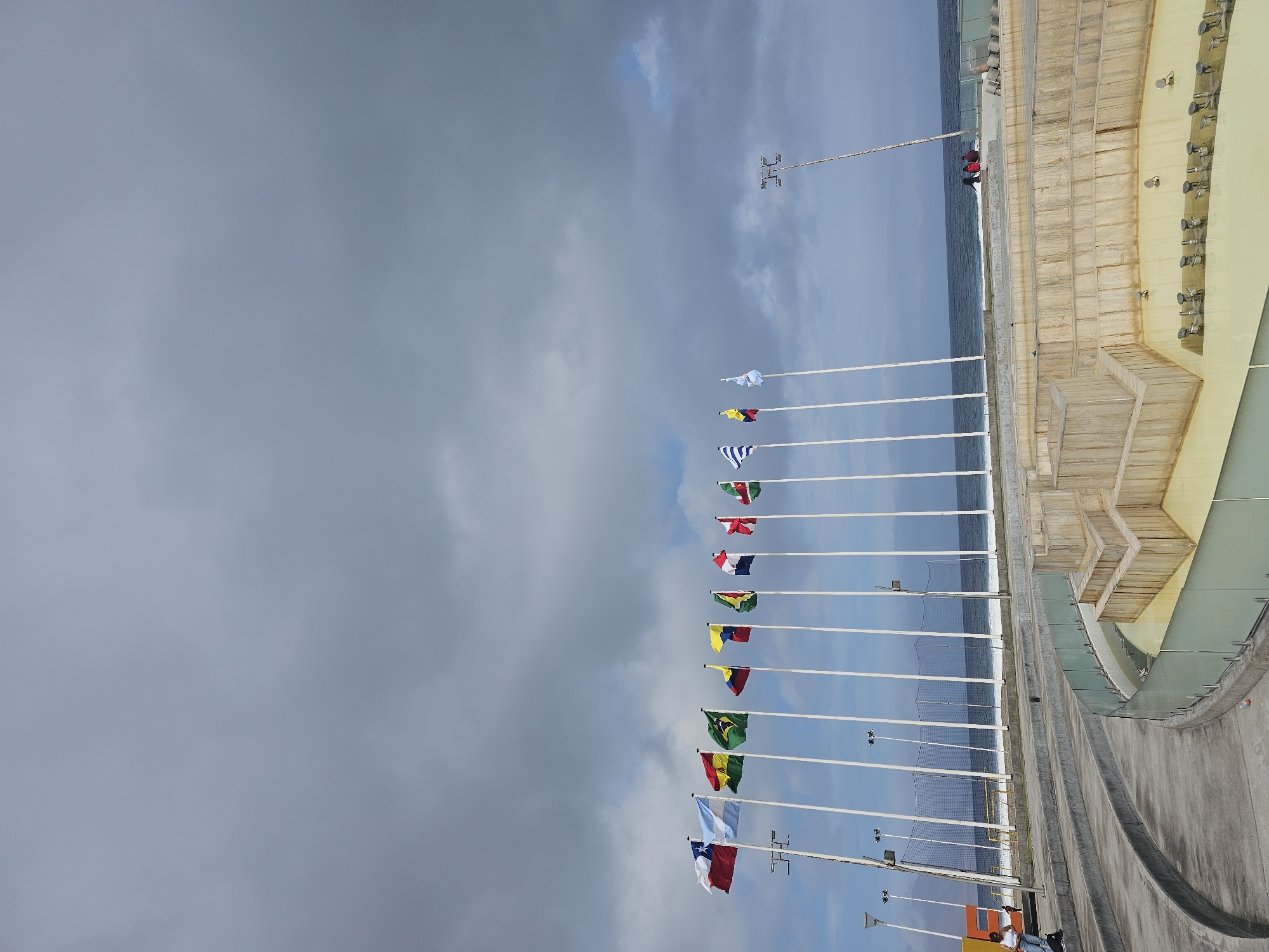The image depicts a row of national flags flying on poles along the coastline in Iquique, Chile. The flags include those of Chile, Argentina, Bolivia, Brazil, Colombia, Ecuador, Paraguay, Peru, Uruguay, and Venezuela. The scene is set against a backdrop of a cloudy sky and the ocean. In the foreground, there is a concrete seating area with a dry fountain in the center. Two individuals are seated on the edge of the fountain, looking out at the sea. The overall atmosphere is calm and suggests a public space near the beach.