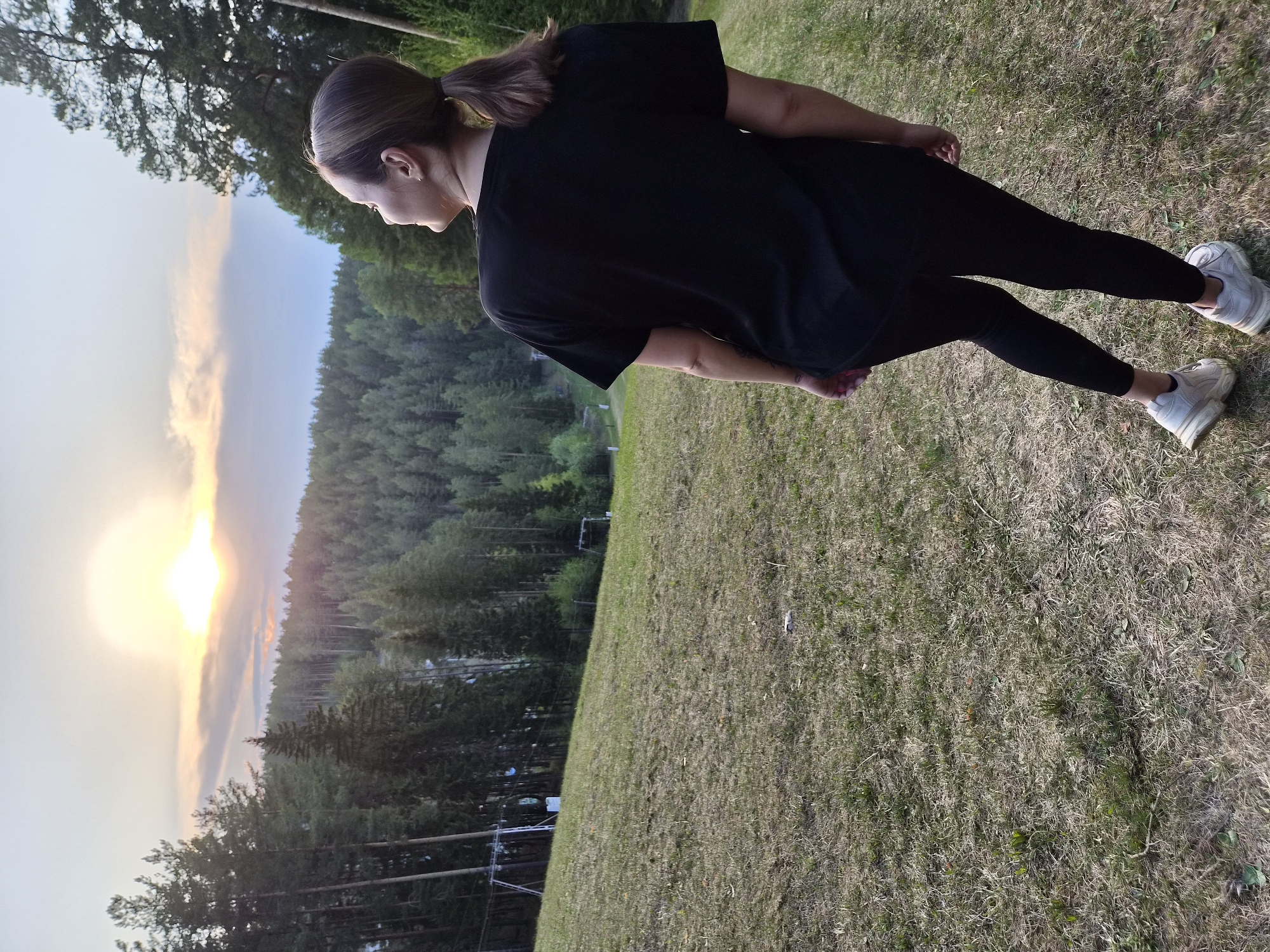 A woman stands on a grassy hill. She faces towards the sky with the sun setting. The woman has her hair in a ponytail and is wearing a black t-shirt, black pants, and white sneakers. There is a forest on the opposite side of the hill.
The location is Прикамский, Russia.
