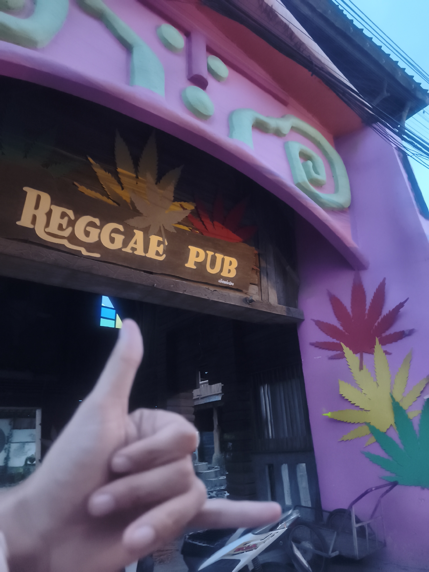 The image depicts the exterior of "Reggae Pub" in Baan Chaweng, Thailand. The entrance is framed by a pink arch decorated with green accents and cannabis leaf designs in red, yellow, and green. The pub's sign, above the doorway, includes the words "REGGAE PUB" above a cannabis leaf. A blurred hand is in the foreground, performing a "shaka" sign. In the background, parts of a building, possible vehicle, street objects, and a glimpse of the sky are visible.