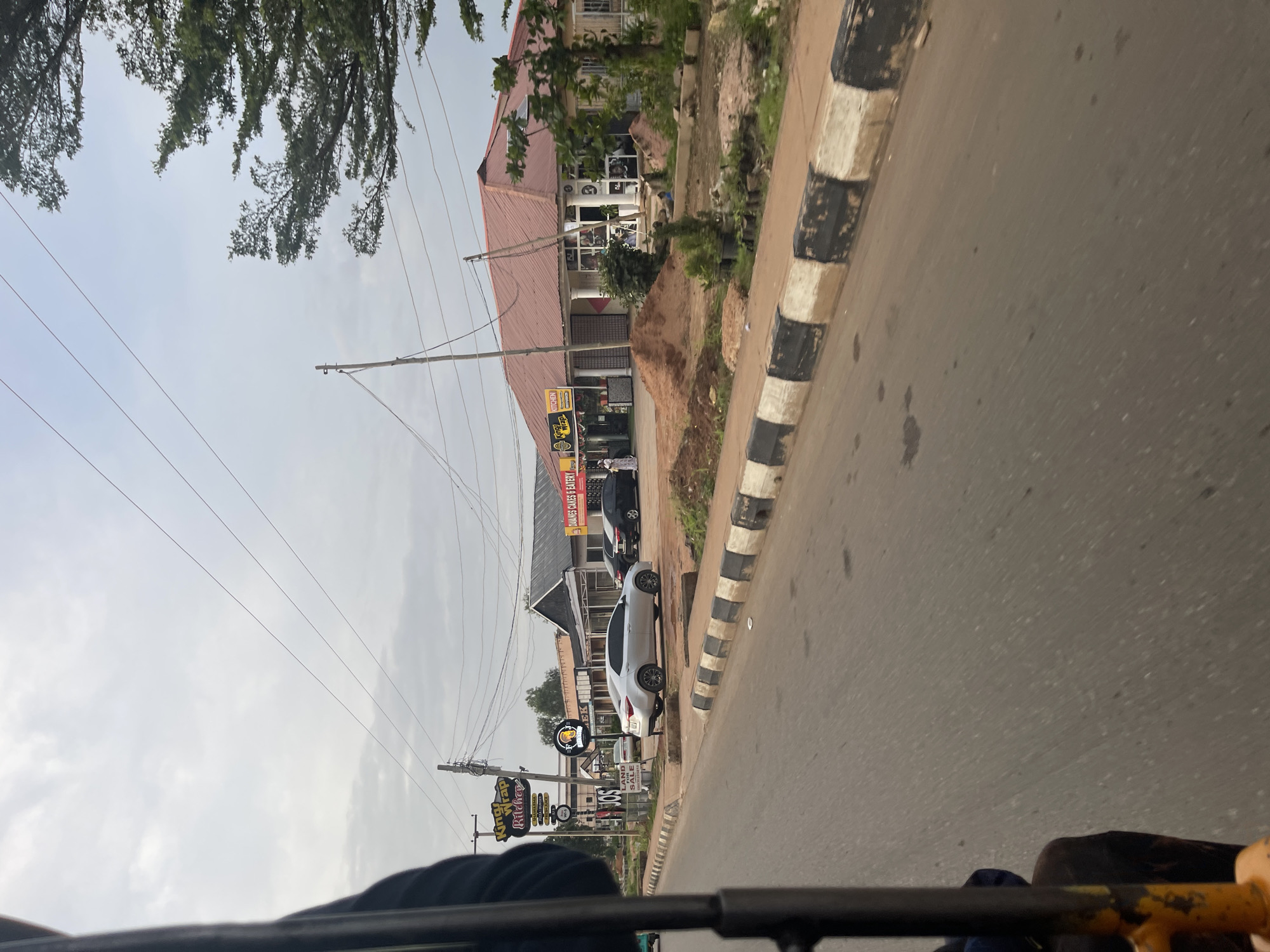 The image captures a street scene in Bar Jirgi Summa, Nigeria, from the perspective of a moving vehicle, likely a rickshaw or a similar mode of transport, as indicated by the partial view of a metal frame and a seat in the foreground. The street is paved, with a distinctive black and white striped curb running along the side. On the right side of the street, a row of buildings with red tiled roofs and various shopfronts is visible. Signs indicate businesses such as "King's Wrap Kitchen" and "DONUTS CAKES & EATERY." Several cars are parked along the roadside, including a silver sedan, a black sedan, and another silver car further down. People are seen walking in front of the shops. The environment appears to be a commercial area with a somewhat overcast sky. Utility poles and power lines crisscross the sky above the buildings. The overall impression is that of a moderately busy street in a Nigerian town.