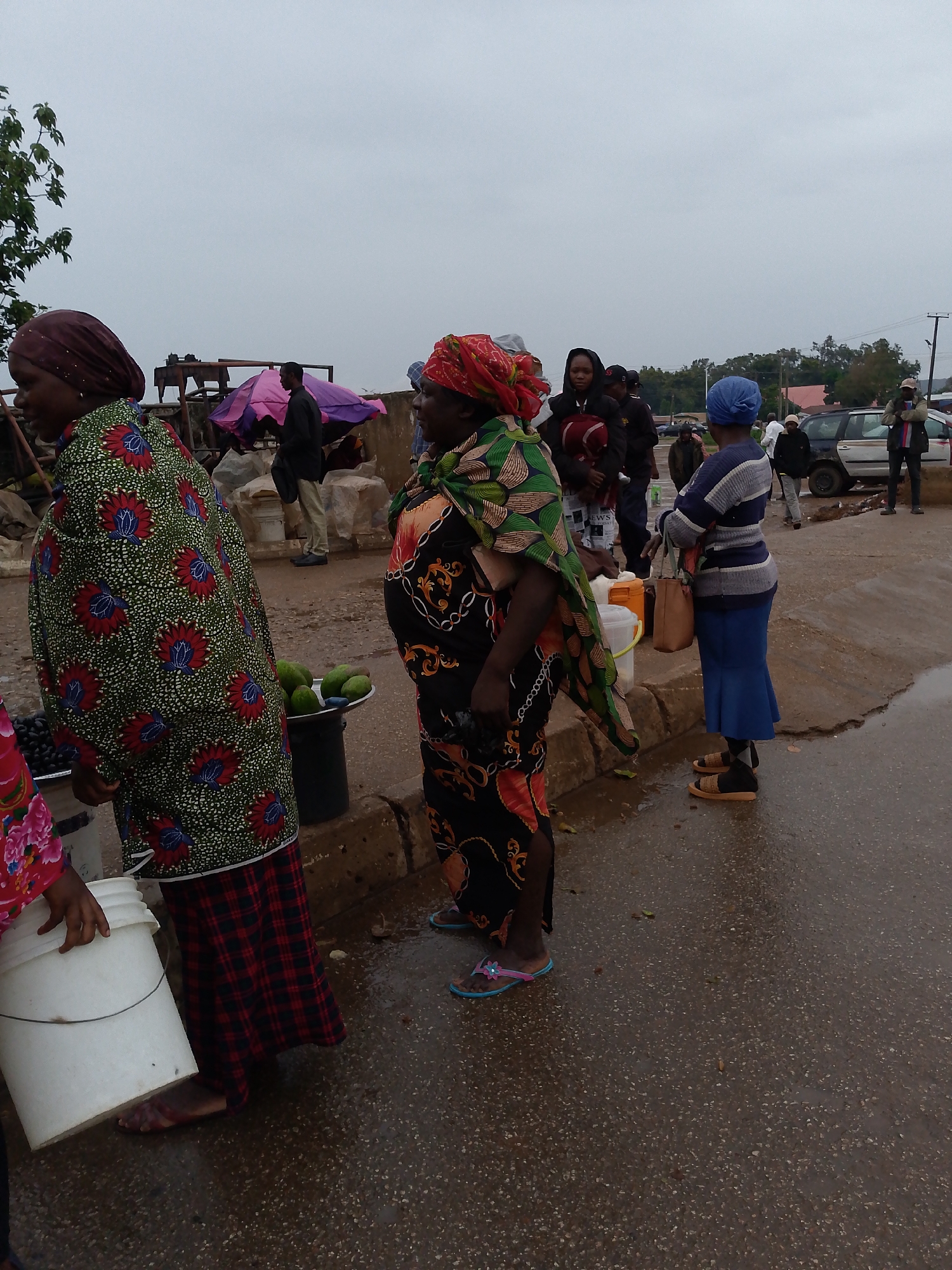 The image depicts an outdoor street scene in Bukuru, Nigeria, on an overcast day with wet ground. Several people, primarily women, are visible, suggesting a busy public area or market.
In the left foreground, a woman wears a maroon headscarf, a green top adorned with red and blue floral patterns, and a red plaid skirt. She holds a white plastic bucket. Beside her, a woman in a black, orange, and gold patterned dress wears a distinctive green and multi-colored patterned headscarf or wrap over her shoulders. She looks towards the right, and her feet in light blue flip-flops are visible on the wet asphalt. Between these two women, a black bucket holds several green fruits, possibly avocados or mangoes, positioned on the curb.
Further into the mid-ground, a woman facing away from the viewer wears a blue headscarf, a blue and white striped long-sleeved top, and a blue skirt, with tan sandals on her feet. She carries a brown handbag. Near her, a person in a dark jacket holds a small bundled object or child, with crumpled paper that partially shows the word "NEWS" in black text. Behind them, a man stands under a pink and purple umbrella.
The background includes other individuals, some men and women, and a silver car parked on the far right. A man in a light-colored hat and jacket stands near the car. The sky is uniformly overcast, and the dark asphalt ground shows numerous puddles and wet patches, indicating recent rain or damp weather conditions. Trees and some distant buildings are visible on the horizon. The scene portrays daily life in a bustling outdoor environment.