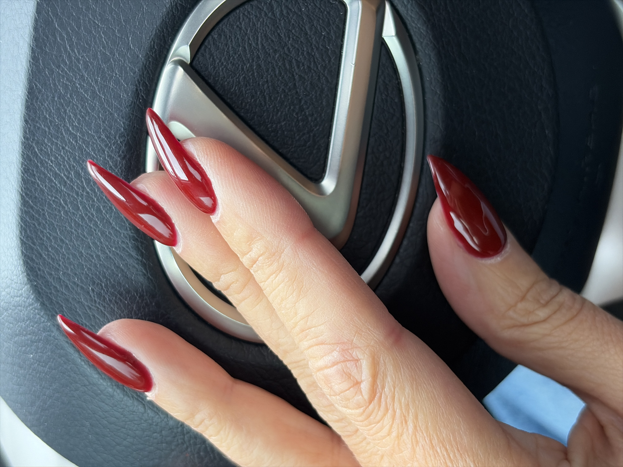 The image shows a close-up of a person's hand with long, stiletto-shaped, dark red nails. The hand is resting on a black leather steering wheel, which features a metallic emblem, likely a car logo. The lighting suggests an outdoor setting, possibly during daylight. No specific location in Balashikha, Russia can be determined from the visual information provided.