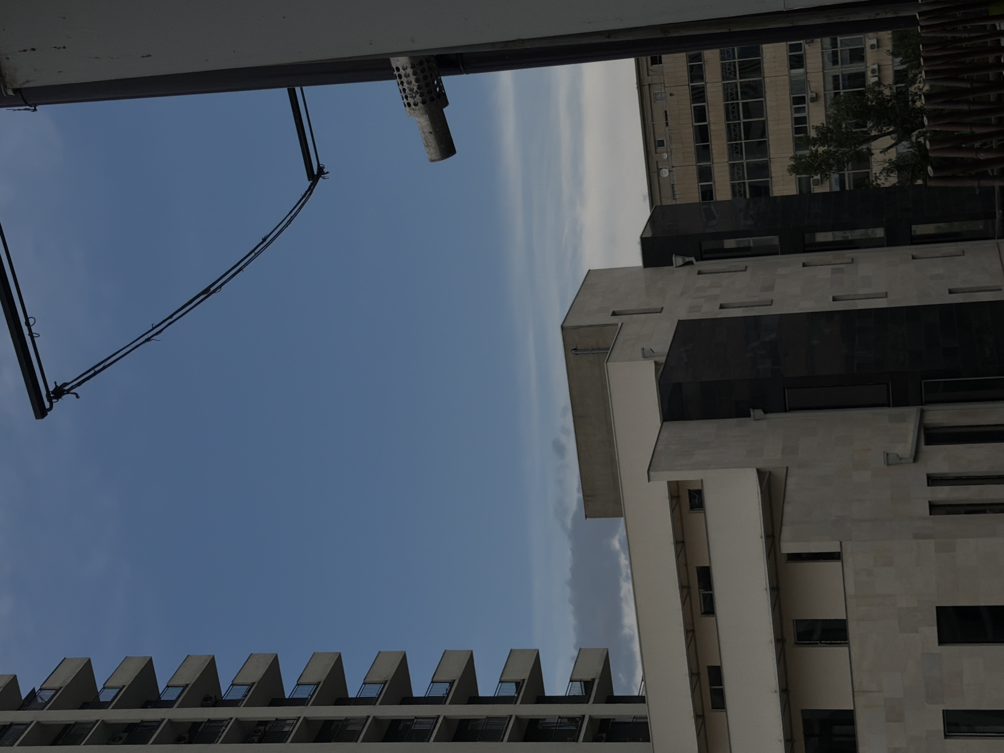 The image depicts a view of buildings in the city of Tbilisi, Georgia. On the left, a tall, modern residential building with numerous balconies is visible against a blue sky. In the center, a lighter-colored building with a stone facade and dark window accents is partially obscured by the residential tower. To the right of this, another building with a dark facade stands. Further in the background, a larger, older building with a grid of windows and visible air conditioning units is present, with trees and greenery in front of it. Overhead, power lines and a support structure are visible, as well as what appears to be a drainage pipe or vent attached to a pole on the far right. The overall impression is of an urban environment with a mix of architectural styles.