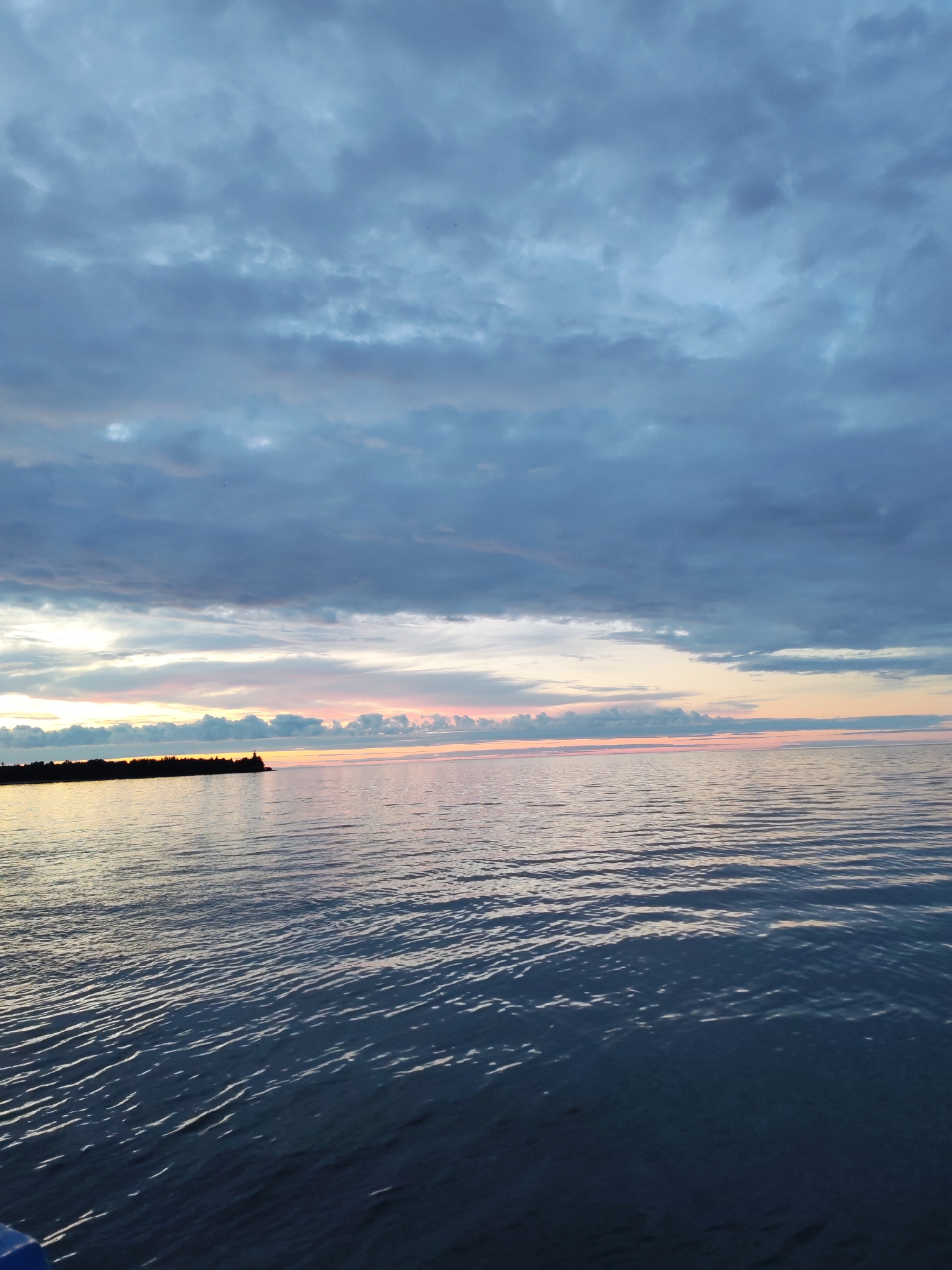 The image depicts a serene seascape at dusk, featuring a body of water reflecting the muted colors of the sky. A dark, silhouetted landmass, possibly a peninsula or a shoreline with a lighthouse, juts out from the left side of the frame. The sky is dominated by dramatic, dark blue and gray clouds, with a sliver of soft pink and yellow light breaking through at the horizon, indicating the setting sun. The water's surface is calm, with gentle ripples creating a textured pattern that catches the faint light. The scene evokes a sense of peaceful isolation. No people or specific urban features of Shcheleiki, Russia are visible in this image.