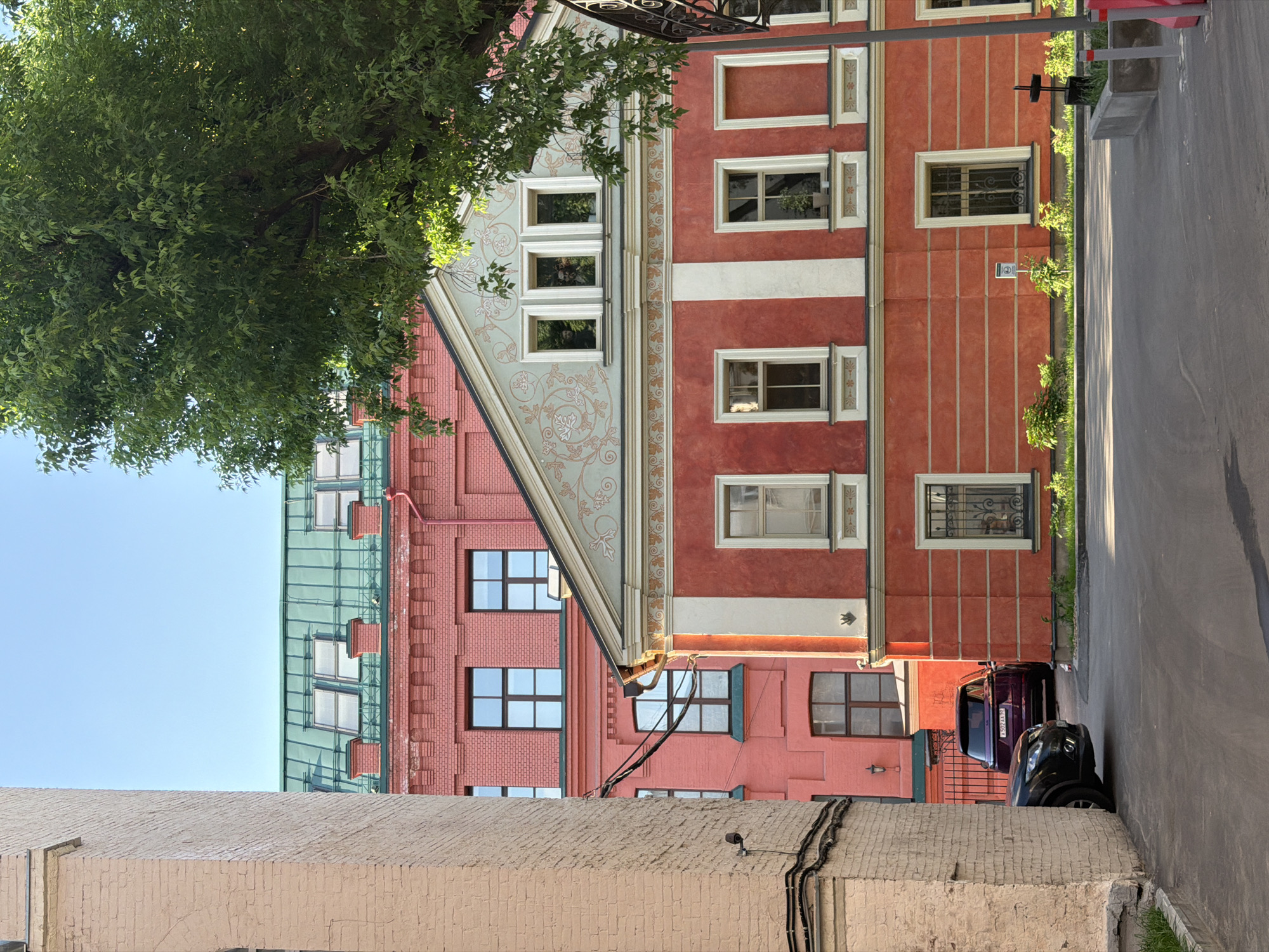 The image shows an urban scene in Moscow, Russia. A portion of colorful buildings in shades of red and peach are visible. The architecture includes patterned and textured facades, multiple windows, and ornate details. The buildings appear to be several stories high. A green tree occupies the left side of the image, partially obscuring the buildings. Below, a light-colored brick wall is seen. A street with parked cars runs along the right side of the image.