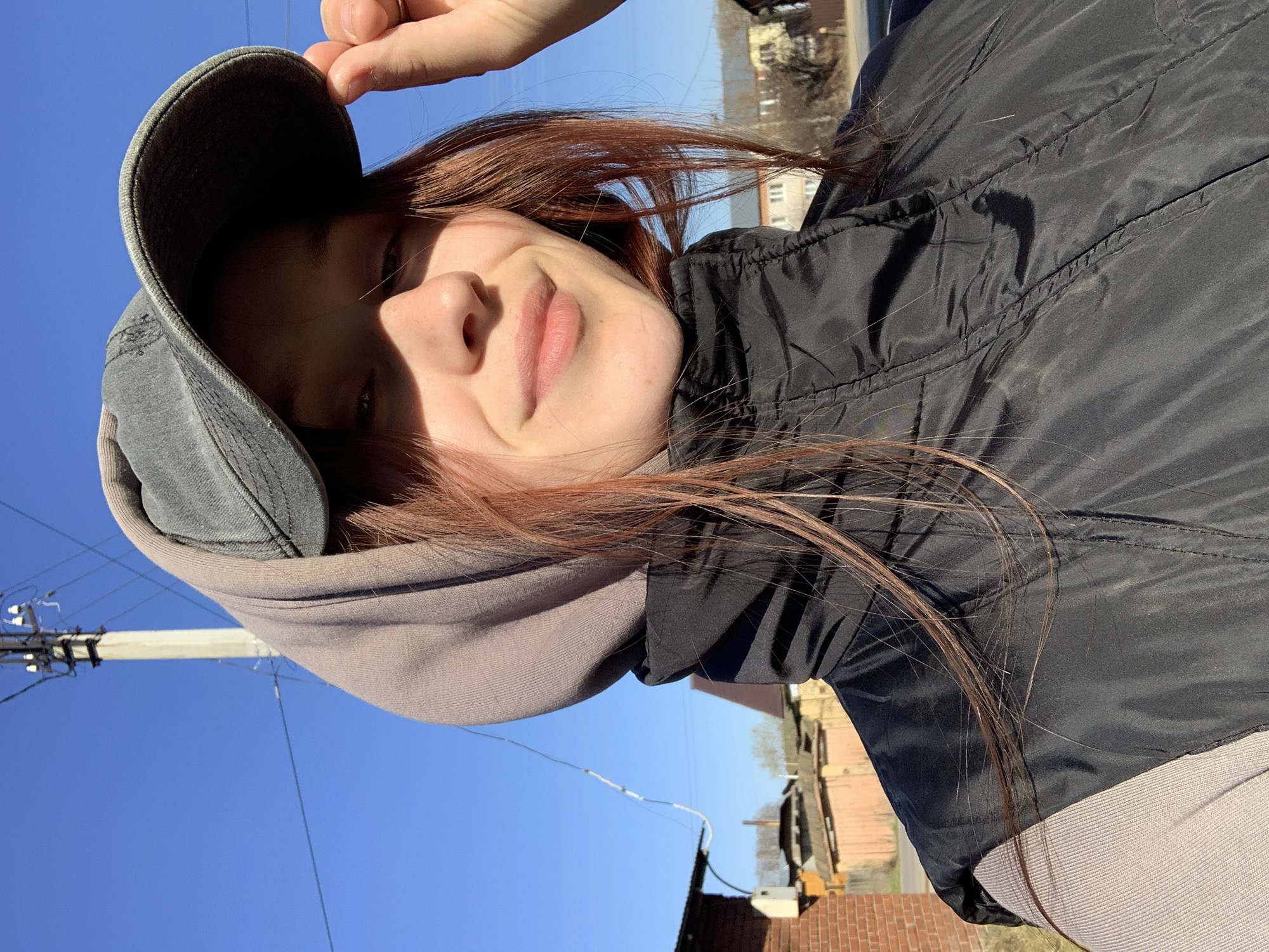 A low-angle, upward-tilted shot captures a young woman in Фоки, Russia. She wears a gray hooded sweatshirt with a dark gray cap pulled low, obscuring part of her face and casting shadows. Her brown hair is visible around the edges of the cap and hood. She is also wearing a puffy black jacket. She holds the edge of her cap with her hand. The background features a clear blue sky, telephone poles and wires, and the roofs of brick buildings.