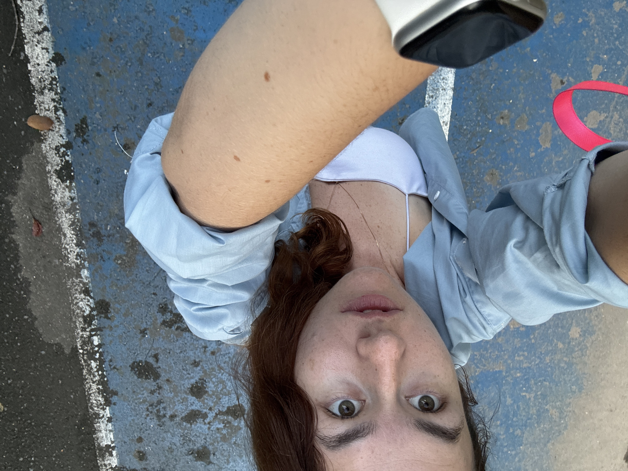 The media file shows a close-up, high-angle selfie of a young woman with reddish-brown hair. She is wearing a light blue, open button-down shirt over a white bikini top and a delicate gold necklace. Her eyes are wide open and looking directly at the camera, conveying a sense of surprise or excitement. She has freckles scattered across her face and light-colored eyebrows.
The setting appears to be outdoors, possibly a public space with a blue, worn-down surface, possibly a basketball court or a parking lot, marked with a white line. There are some dark, damp patches on the ground, suggesting it might have rained recently or the area is wet. A bright pink leash or strap is visible in the upper right corner of the image. The lighting suggests it is daytime, with natural light illuminating the scene. The overall impression is a candid and spontaneous moment captured during an outing. The location context provided is Phuket City Municipality, Thailand.