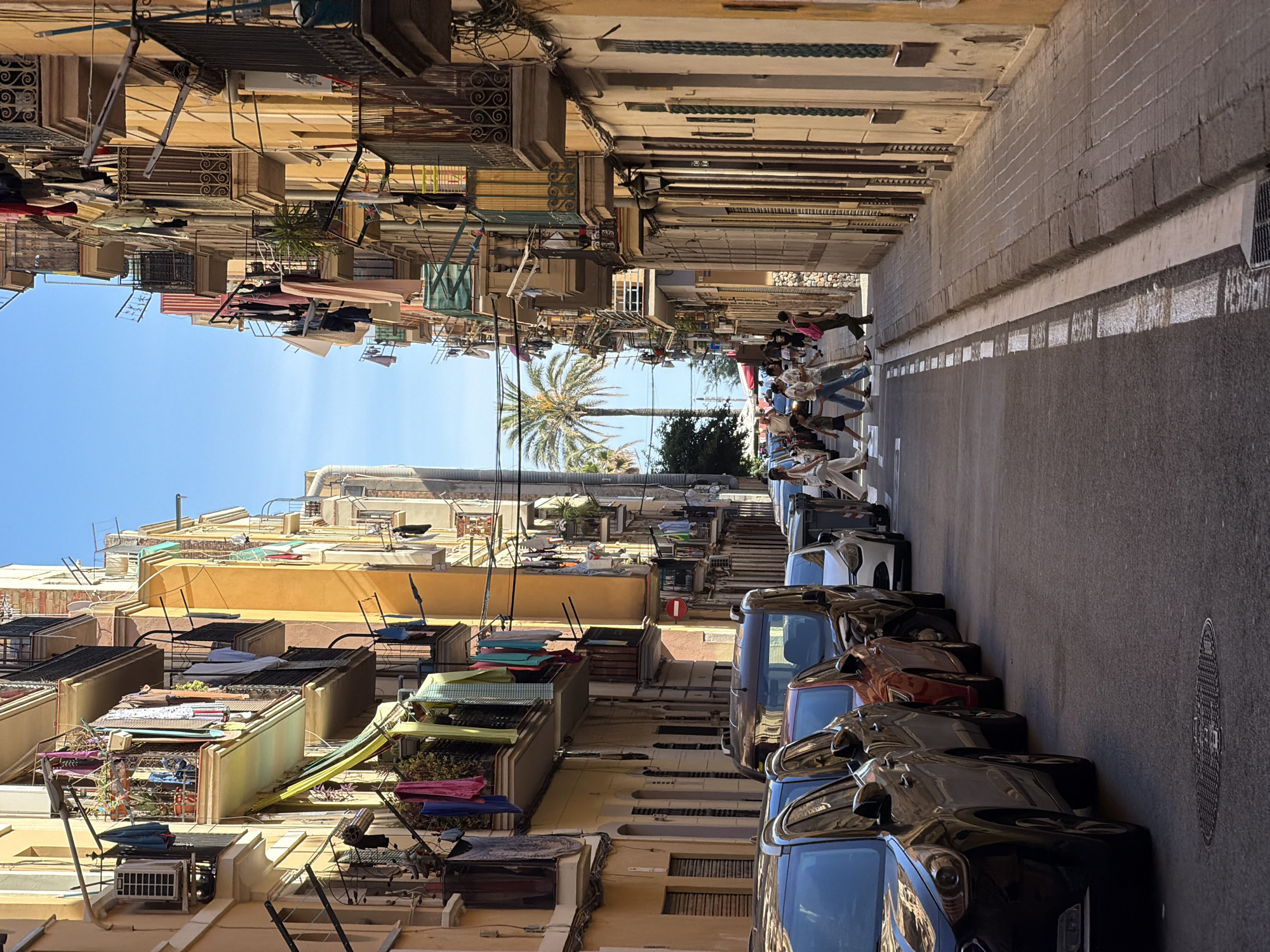 This image captures a sun-drenched, narrow street in the Gothic Quarter of Barcelona, Spain. The architecture features tall, colorful buildings with balconies adorned with drying laundry, a quintessential sight in this historic district. A group of people, casually dressed, are crossing the street at a designated crosswalk. The street is lined with parked cars, and a palm tree is visible in the distance, adding to the Mediterranean ambiance. The sky is a clear blue, indicating a warm and sunny day. The overall scene conveys a sense of everyday life in a vibrant European city.