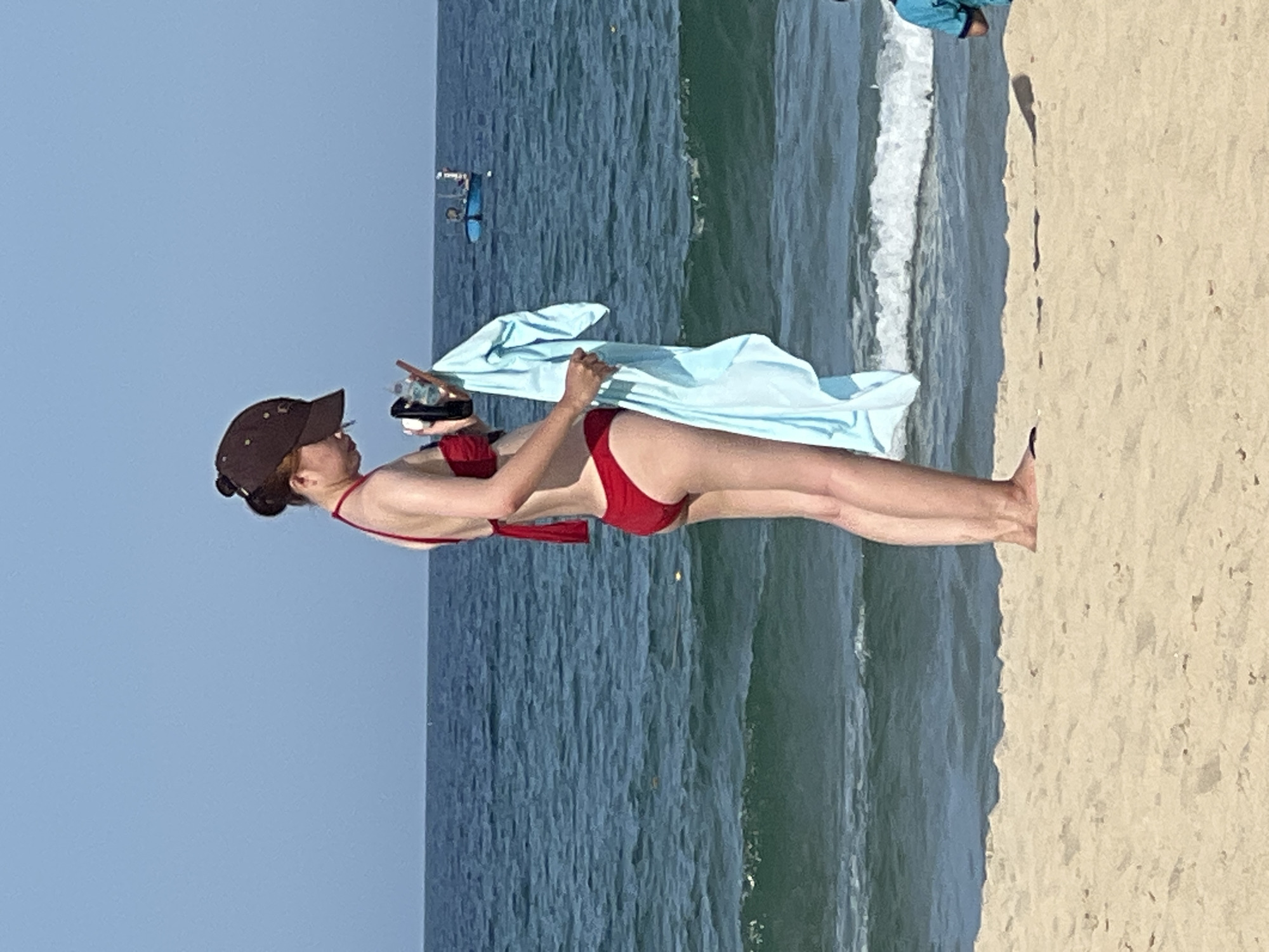 This image captures a scene at a beach in Đà Nẵng, Vietnam, under a clear blue sky. In the foreground, a woman wearing a red bikini and a brown baseball cap stands on the sand, facing the ocean. She is holding a light blue towel and a black object, possibly a phone or camera, in her hands. Her posture suggests she is looking at the object. In the mid-ground, the ocean stretches out, with gentle waves breaking near the shore. A small blue boat with two figures on board is visible in the distance on the water. In the far right of the image, the legs of a person in a blue shirt can be seen standing on the sand. The overall impression is a typical beach scene with elements of recreation and leisure.