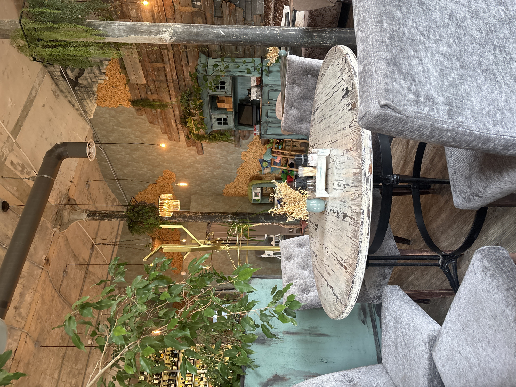 The image depicts the interior of a restaurant in Ivanovo, Russia, featuring a rustic and eclectic design. A round, distressed wooden table is centrally located, surrounded by four upholstered grey chairs. On the table are salt and pepper shakers, a small vase with dried flowers, and a small white tray.
The background reveals a variety of textures and decorative elements. To the left, a large leafy plant cascades from above, partially obscuring a turquoise bar or counter displaying rows of bottles. A tall, gold coat rack stands behind the plant.
The walls are a mix of textures, with some areas appearing to be rough plaster or concrete, while others are covered with stacked wooden planks. Decorative elements on the walls include clusters of yellow material and hanging plants. A vintage-style turquoise cabinet with open shelves and glass-fronted upper cabinets is visible, holding various items including a computer monitor and baskets.
Industrial elements are also present, such as exposed pipes and a concrete ceiling with visible conduits. Lighting fixtures include pendant bulbs and a crystal chandelier. The overall atmosphere is warm and inviting, with a bohemian or industrial-chic aesthetic. No people are visible in the image.