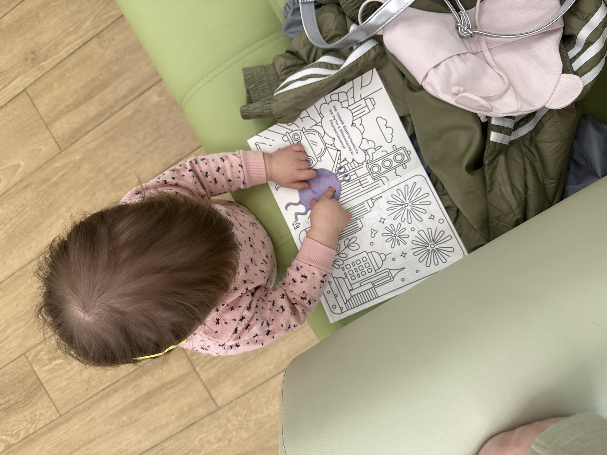 A high-angle shot shows a baby with brown hair, wearing a long-sleeved pink shirt with black floral patterns, sitting on a light green chair. The baby is coloring on a piece of paper with a purple crayon. The paper has a black-and-white drawing of buildings, fireworks, and transportation vehicles with text in Russian which translates to "Do not lose your vigilance on the way to the Moscow Polyclinic!". The paper is placed on a pile of clothes, including a green jacket with white stripes, a pink hoodie, and a silver strap. A light-colored wooden floor is visible in the background. The location depicted is Moscow, Russia, as indicated by the Russian text mentioning "Moscow Polyclinic."
