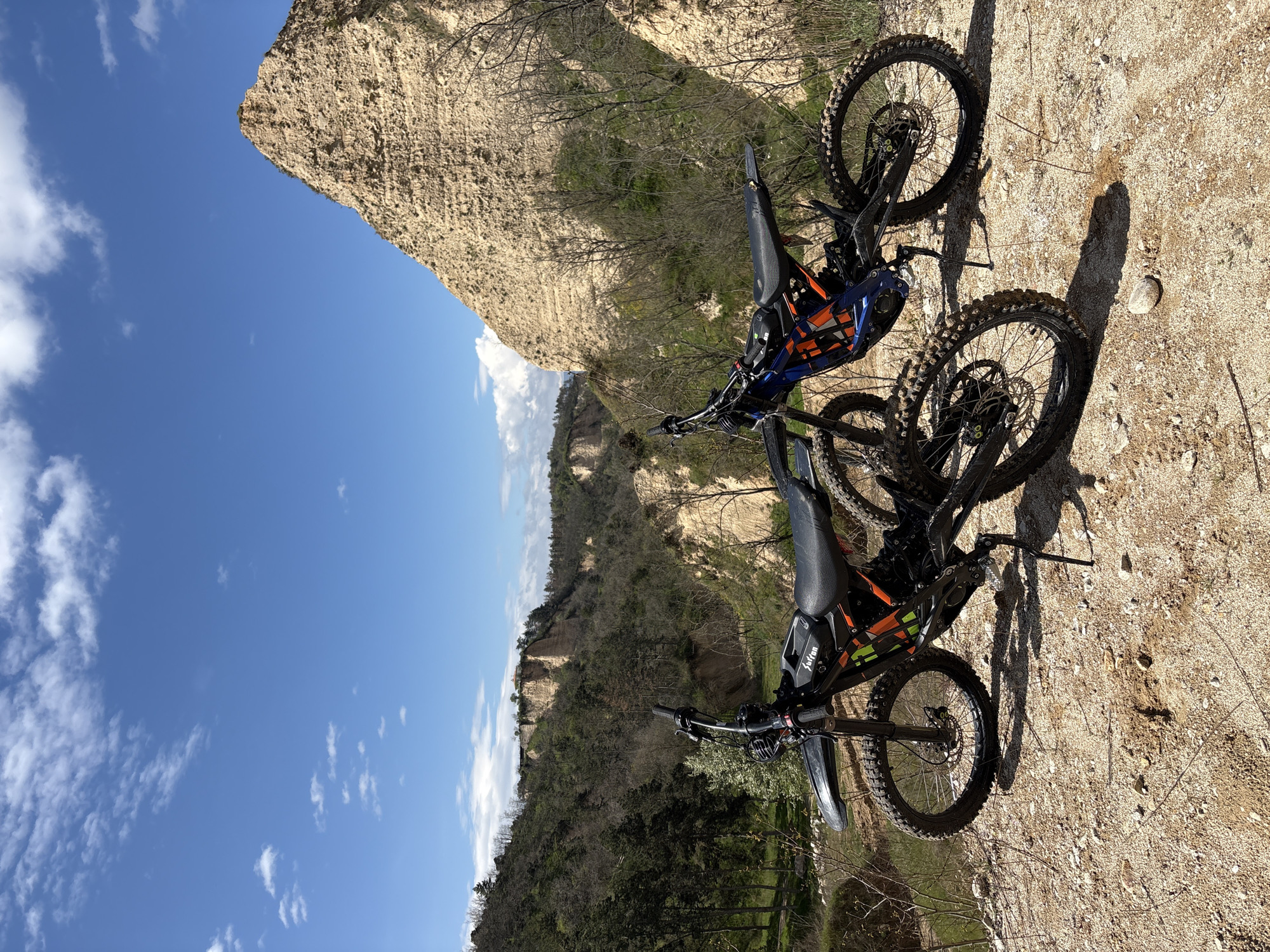 Two Sur Ron electric dirt bikes are parked on a dirt path near the sandstone cliffs of Melnik, Bulgaria. The sky is blue with scattered white clouds. Green vegetation and trees are visible along the hillside.