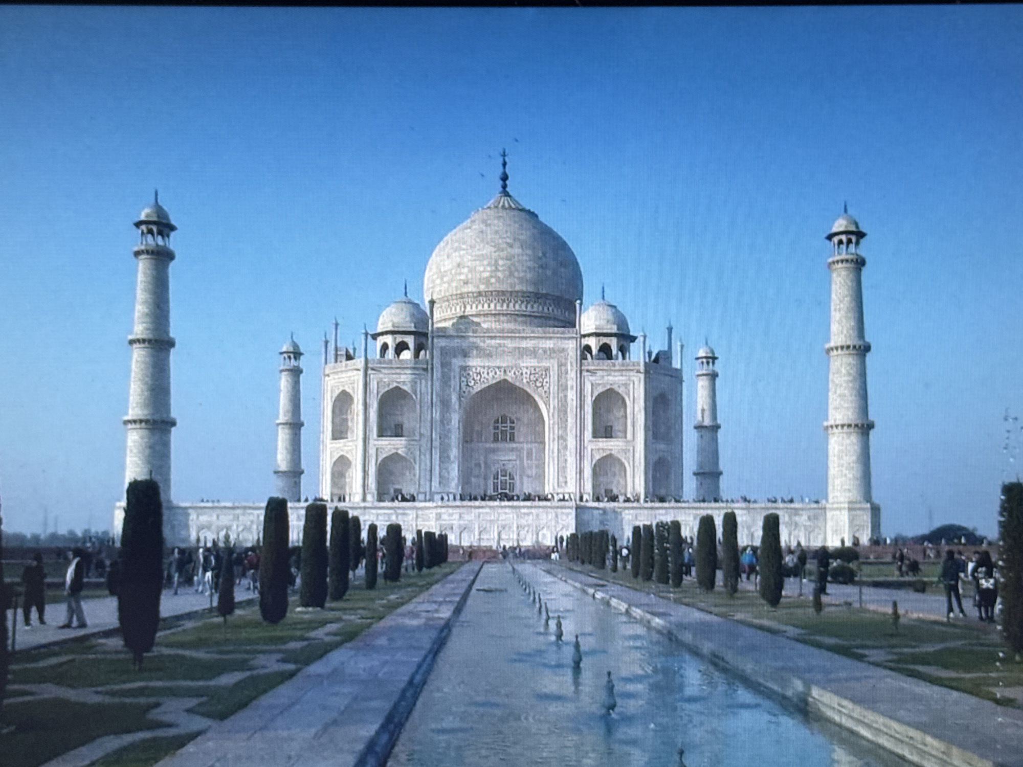 The image depicts the Taj Mahal, a white marble mausoleum located in Agra, India. The iconic structure is centrally positioned, with its dome and minarets prominently visible against a clear blue sky. A reflecting pool runs down the center of the foreground, with water fountains sparsely distributed. Tall, dark green cypress trees line the pathways on either side of the pool, leading towards the mausoleum. Several people are scattered throughout the scene, walking along the pathways and standing near the Taj Mahal. The overall environment appears to be a park or garden setting.