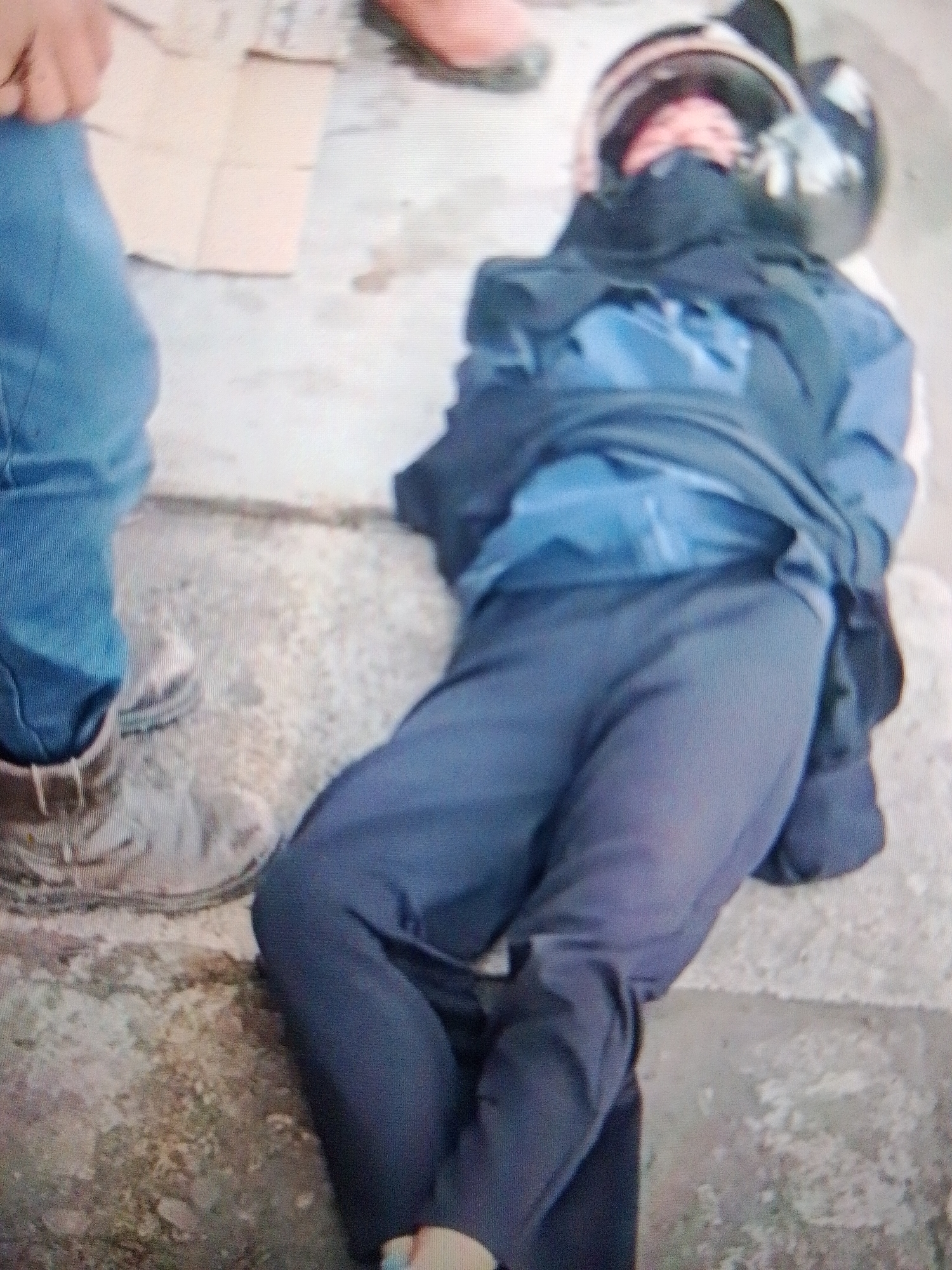 The image shows a person lying on the ground, wearing a helmet, dark clothing, and blue pants. They appear to be an adult male, but their face is partially obscured by the helmet and clothing. The person is lying on their back with their legs bent and crossed. Their right arm is bent at the elbow, and their left arm is not visible. The person's body is covered by a dark blue jacket or coat, which is partially unzipped, revealing a lighter blue shirt underneath.
The setting appears to be outdoors, on a rough, concrete surface with some dirt and debris. To the left of the person, there are the legs of another individual wearing blue jeans and brown shoes. In the background, a piece of cardboard is visible on the ground.
The overall impression is that of an accident or incident, possibly involving a motorcycle, given the helmet. The lighting suggests it could be daytime, but it's difficult to ascertain the exact time. There are no clear indications of weather conditions. The scene lacks any strong emotional cues, and no text is visible in the image. The location context provided is Madukoro, Indonesia, which may offer further clues to the specific circumstances of the image.