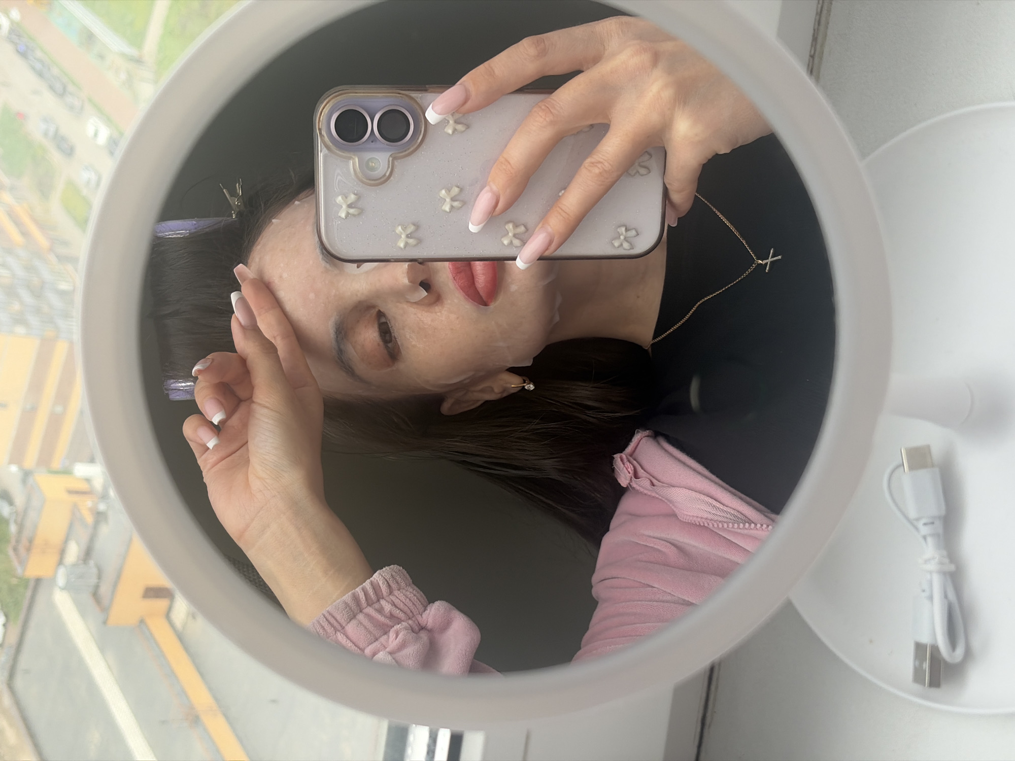 A reflected image shows a person taking a selfie in front of a round vanity mirror. The person is wearing a sheet face mask and holding a mobile phone, its case decorated with small white floral embellishments. The phone's dual camera setup is visible. The person's nails are manicured with a French tip design. A piece of purple hair accessory is clipped to their hair, with dark brown hair strands visible in the mirror. The person is wearing a black top and a light pink jacket. A gold necklace with a cross pendant adorns their neck. The mirror is on a white stand, and a coiled white USB charging cable is on the mirror's base. In the blurry background, one sees the window that offers a glimpse of the city outside with yellow-beige buildings and other architectural structures under an overcast sky.