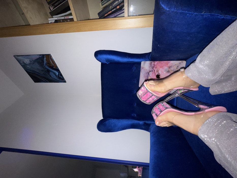 The image displays an interior scene from a low-angle perspective, focusing on a person's lower legs and feet. The individual is seated in a royal blue velvet armchair, extending their legs. They are wearing shimmering silver trousers and clear, open-toed platform high-heeled sandals with pink insoles and transparent stiletto heels. A small electronic device, possibly a tablet or laptop, with a pink marble-patterned case and an Apple logo, rests on the armchair next to their feet.
The background features white walls. To the right, a light wooden bookshelf is visible, populated with various books, some displaying Cyrillic titles on their spines. This detail indicates a Russian-speaking environment. Above the bookshelf, an abstract square painting with swirling patterns in shades of blue, white, and brown is mounted on the wall. A vertical royal blue structural or furniture element is present on the far left. Specific identification of the location as Moscow, Russia, is not ascertainable from the visual information beyond the inference of a Russian-speaking country.