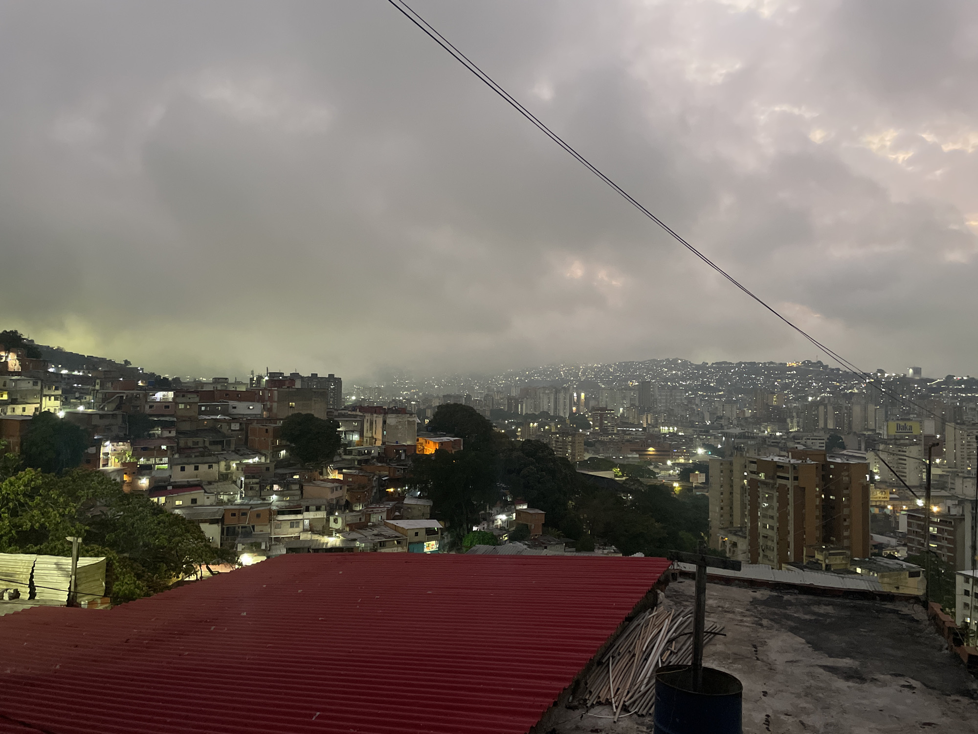 The image depicts an overcast evening cityscape, likely Caracas, Venezuela, given the high-density urban environment and mountainous terrain. The foreground features a red corrugated metal roof and a flat concrete surface with debris, including wood planks and a blue barrel. Beyond the roof, a densely packed hillside community with numerous closely-built houses extends toward the left side of the frame. Streetlights and illuminated windows suggest early evening. In the middle ground, a valley contains dense trees and vegetation. The background shows a sprawling cityscape with numerous high-rise buildings illuminated against the cloudy sky. Overhead power lines traverse the upper portion of the frame.