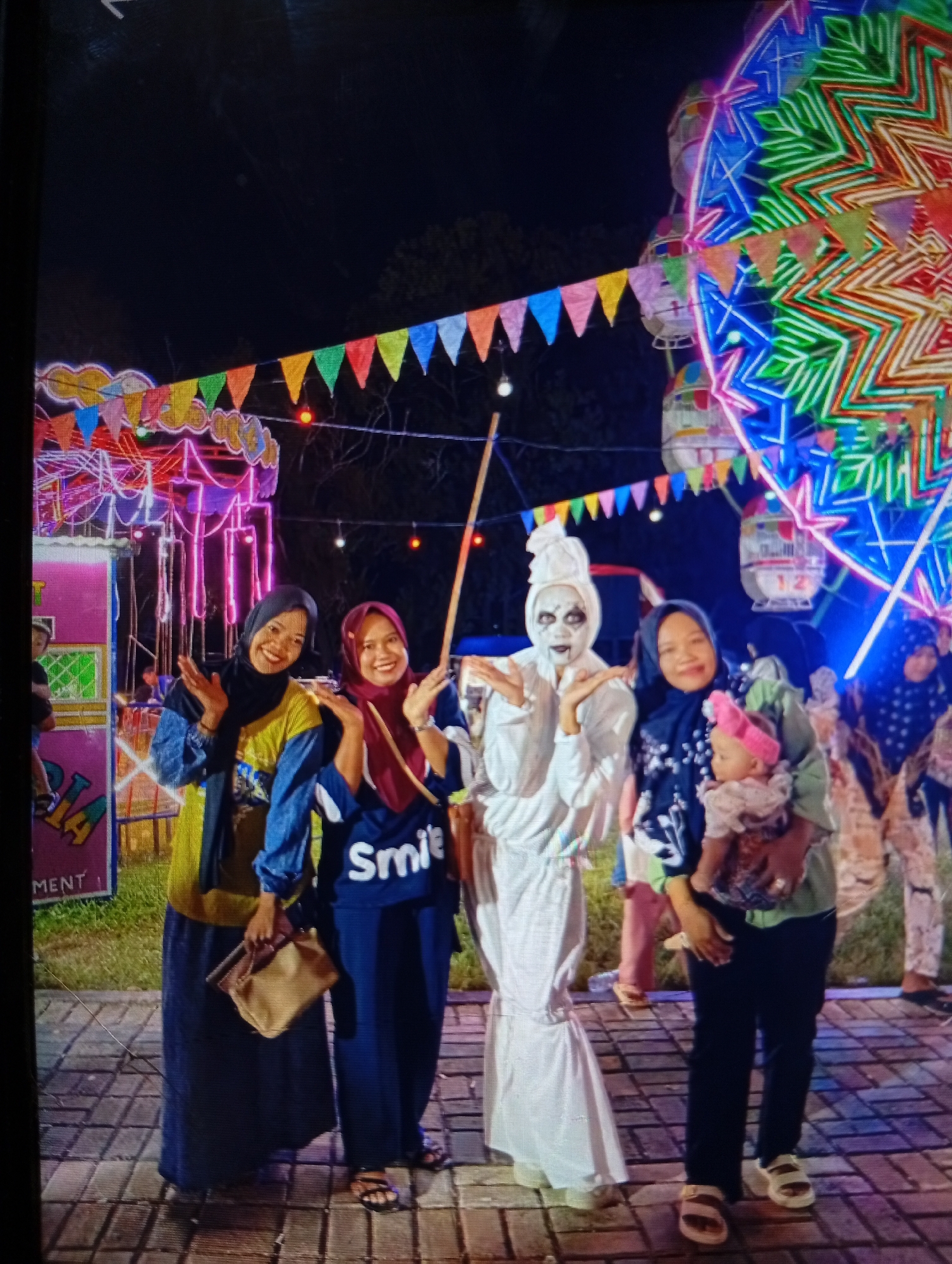 The image captures a nighttime scene at a fairground, likely located in Lhokseumawe, Indonesia, indicated by the presence of colorful carnival lights, decorative flags, and what appears to be a Ferris wheel. Four women are the central subjects. Two women on the left are smiling and posing with open hands. The woman in the center is dressed in a white, ghostly costume with face paint resembling a skull and dark eyes. To her right, another woman holds a baby. The background features vibrant, illuminated structures and a checkered pavement.