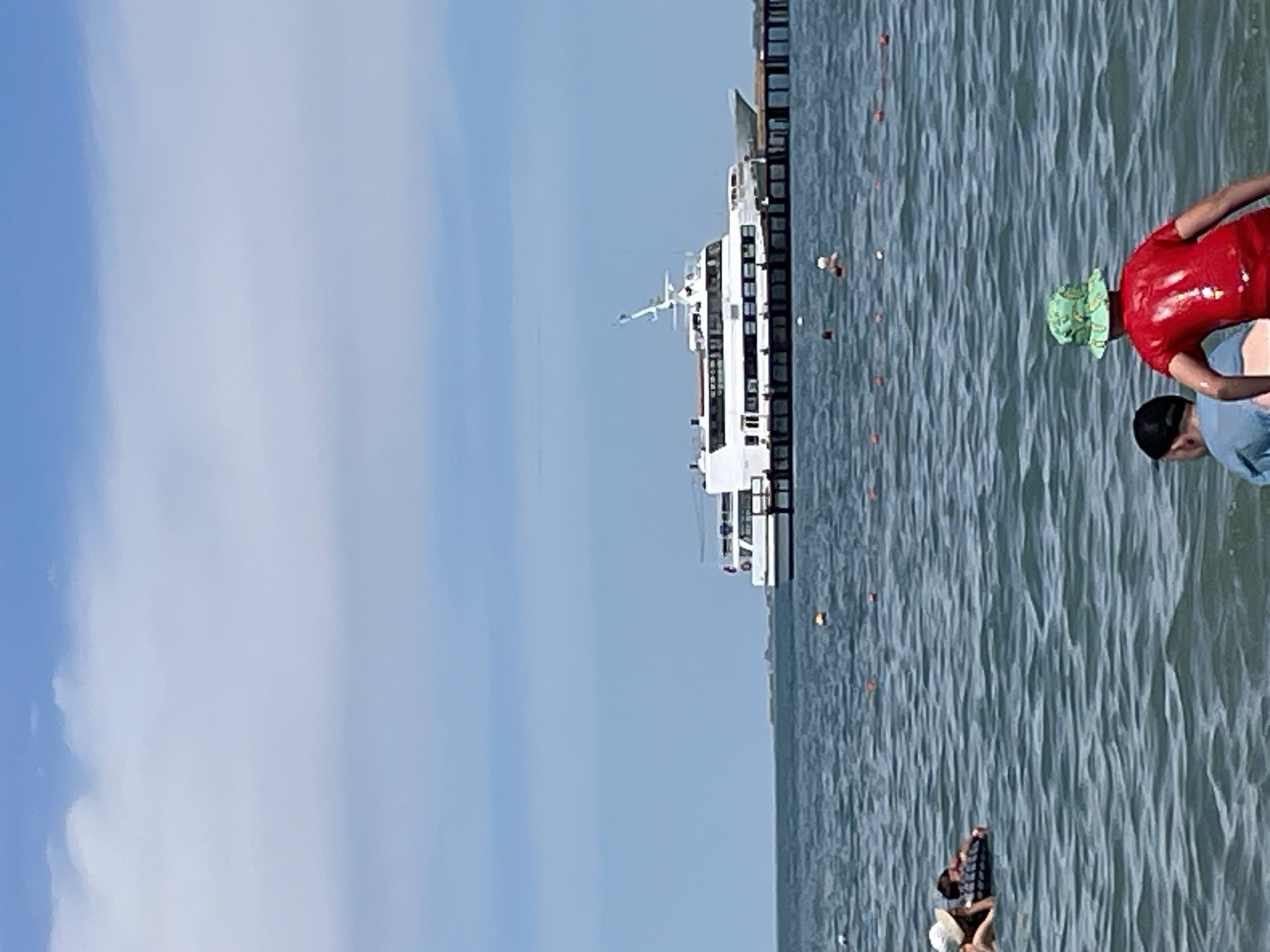 In the city of Unknown, Unknown, a bright, sunny day unfolds at a beach or waterfront. The sky is a vibrant blue, dotted with fluffy white clouds, creating a picturesque backdrop. The water is a deep blue-green, with gentle waves rippling across its surface, indicating a calm and inviting atmosphere.
In the distance, a large, white passenger ferry is docked at a pier, its sleek design hinting at a leisurely journey. The ferry, with its multi-level decks and numerous windows, appears to be a popular mode of transport, ready to welcome passengers for an excursion. The pier, a sturdy wooden structure, extends from the shore, providing access to the ferry and a vantage point for observing the surroundings.
Closer to the shore, several people are enjoying the water. In the foreground, two children, one wearing a red shirt and a green patterned hat, and another in a blue shirt, are seen from behind, facing the water. Their backs are turned to the camera, suggesting they are about to enter the water or are already playing near the shore. A few other individuals can be spotted in the water, some possibly swimming or wading.
The overall scene conveys a sense of relaxation and enjoyment, with the ferry and the presence of people in the water suggesting a popular tourist destination or a local recreational spot. The clear sky and calm water contribute to a pleasant and inviting ambiance.