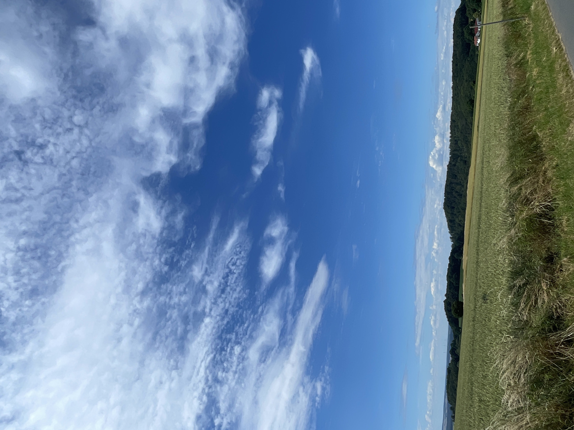 An outdoor, vertical shot captures a vast expanse of a bright blue sky filled with wispy white clouds. Below, a verdant landscape unfolds with rolling hills covered in dense green forest. In the foreground, a field of tall, golden grass sways gently, bordering a cultivated field of light green crops that stretches towards the distant tree line. A small white house with a red roof is visible nestled among the trees on the right side of the frame. The scene depicts a serene rural setting, likely in Christerode, Germany, under a clear, cloud-dotted sky.