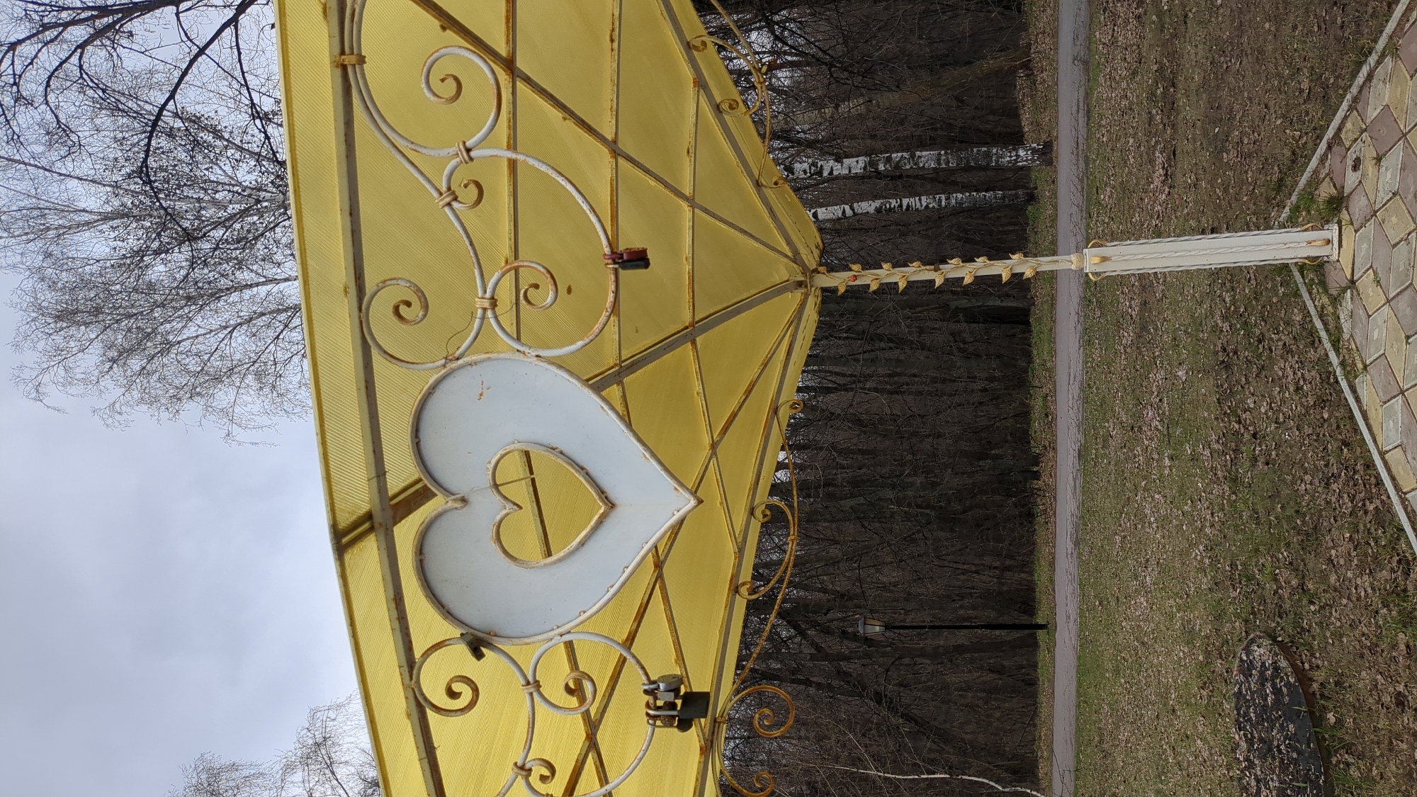 The image captures a decorative yellow canopy or gazebo structure in an outdoor park setting in Ulyanovsk, Russia. The canopy consists of translucent, ribbed yellow material, likely polycarbonate, supported by an ornate framework of white and gold-painted metalwork. A prominent feature is a large white heart-shaped ornament with a smaller heart cutout, centrally located. Intricate white scrolled ironwork embellishes the structure, and several small metal "love locks" are visible attached to the framework.
On the right side, a white support pole, adorned with delicate gold leaf-like decorations, rises from a patterned pavement made of square and diamond-shaped tiles in various shades of brown and tan. The ground surrounding the structure and extending into the mid-ground is covered with sparse grass and a scattering of fallen brown leaves, suggesting a late autumn, winter, or early spring season.
A paved pathway runs horizontally across the scene, separating the immediate foreground from a dense line of bare trees in the background. The sky, visible on the left and above the canopy, is overcast and light gray, with the silhouettes of bare tree branches against it. The lighting is soft and diffused, characteristic of a cloudy daytime. No people or legible text are present in the image.