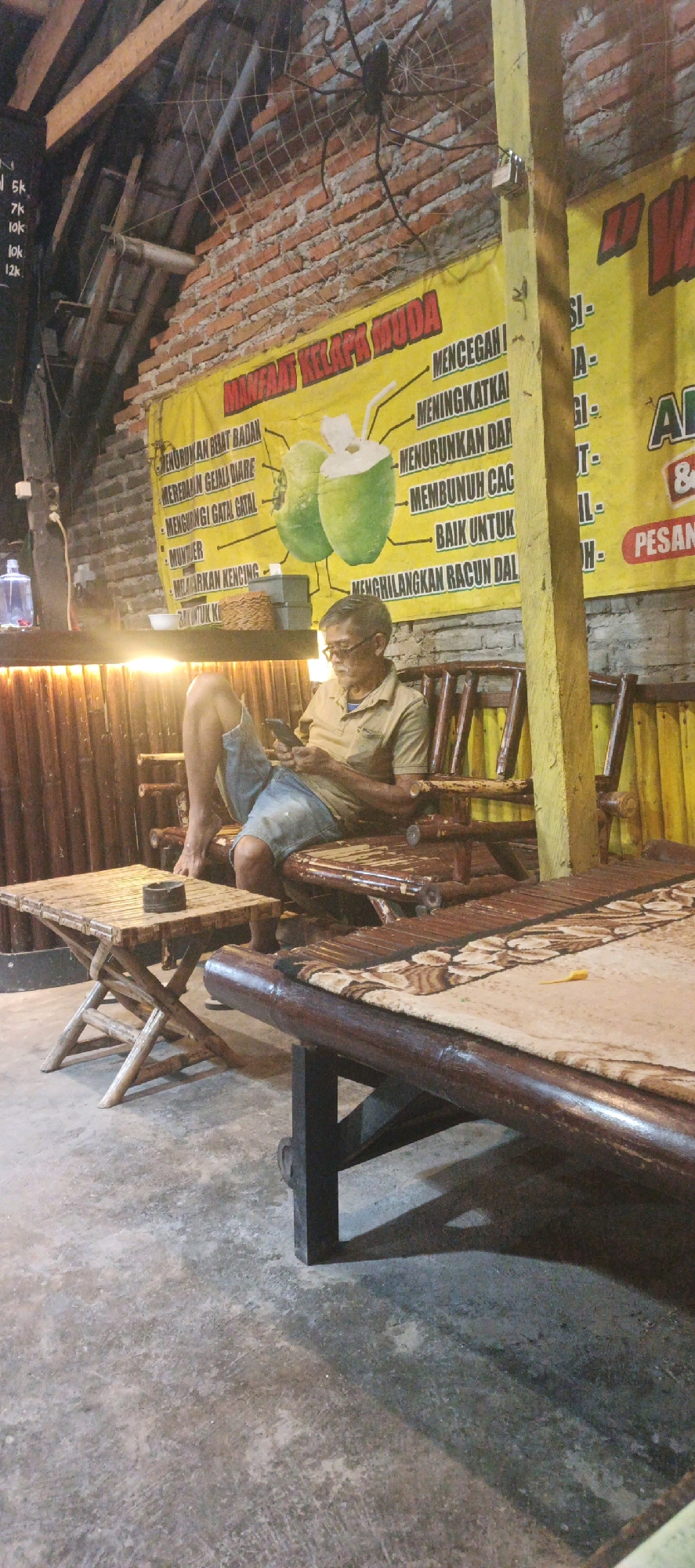 An outdoor, dimly lit seating area in Kasiyan, Indonesia, features a man sitting on a bamboo bench, looking at his phone. He is wearing a light brown short-sleeved shirt and blue shorts. To his left is a small, foldable bamboo table with an ashtray. Behind him, a yellow banner with Indonesian text and an illustration of coconuts is mounted on a brick wall. The structure appears to be a casual eatery or rest stop, with bamboo elements dominating the furniture and decor. The lighting suggests it is evening.